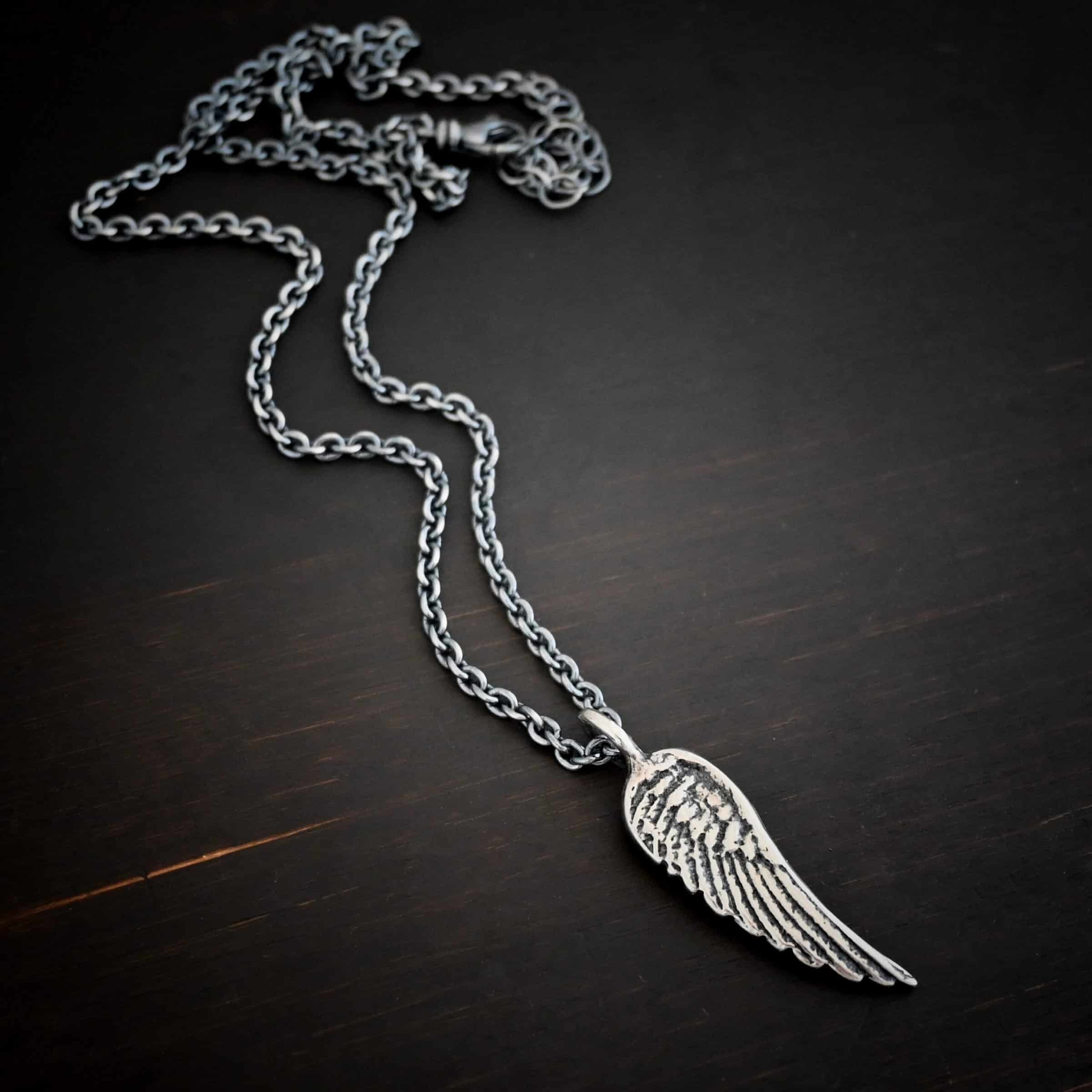 Angel Kiss Necklace Photo of the "Angel Kiss" Necklace. Made out of sterling silver. There is a medium sized angel wing pendant with a loop soldered to the top. There is a chain going through the charm. Necklace is lying against a dark background.