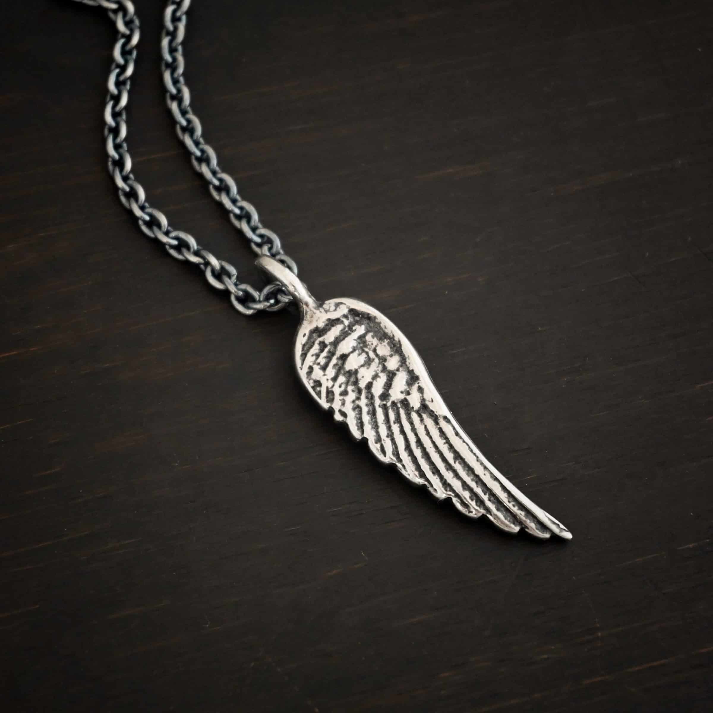 Angel Kiss Necklace Photo of the "Angel Kiss" Necklace. Made out of sterling silver. There is a medium sized angel wing pendant with a loop soldered to the top. There is a chain going through the charm. Necklace is lying against a dark background.