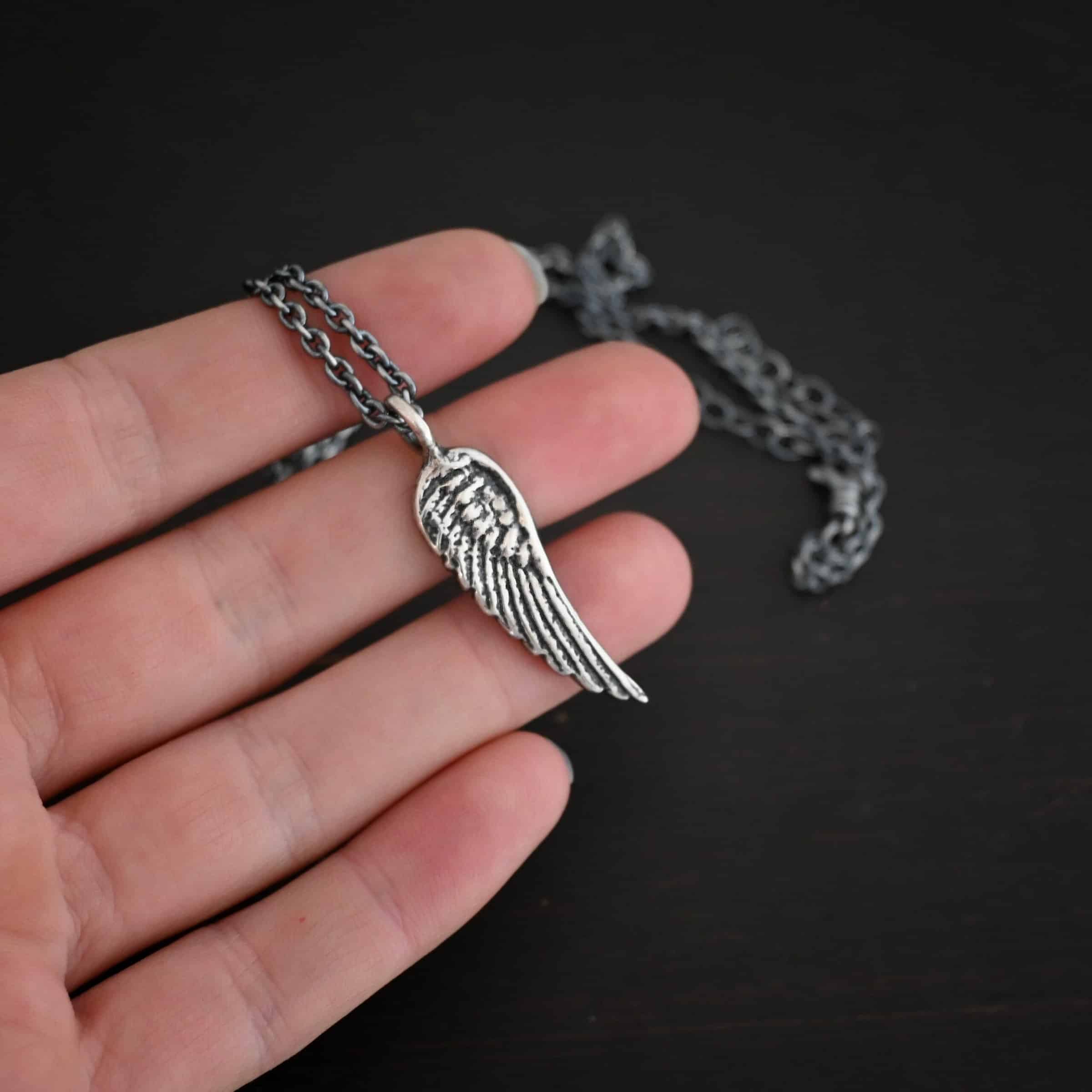 Angel Kiss Necklace Photo of the "Angel Kiss" Necklace. Made out of sterling silver. There is a medium sized angel wing pendant with a loop soldered to the top. There is a chain going through the charm. Necklace is lying against the fingertips of a hand.