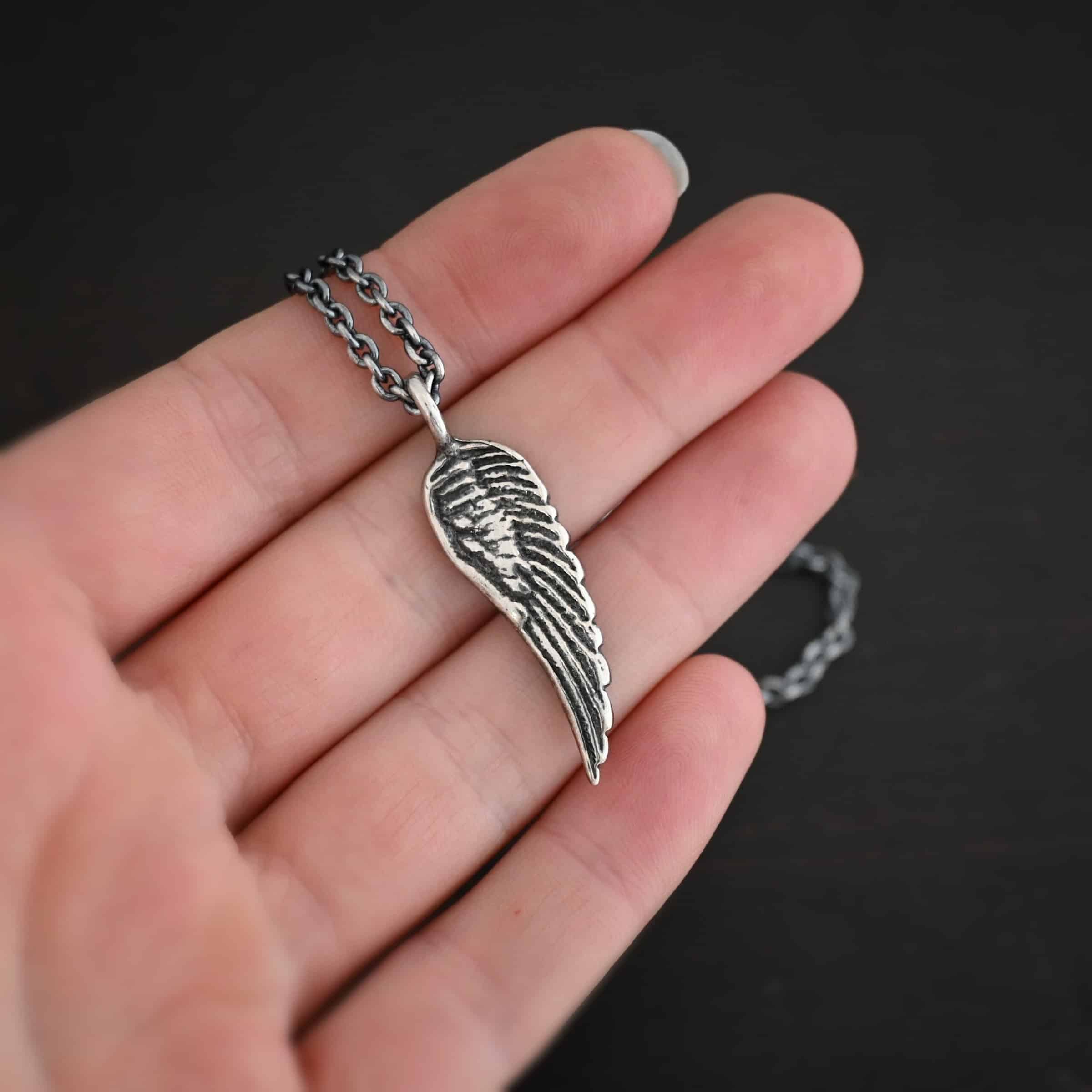 Angel Kiss Necklace Photo of the "Angel Kiss" Necklace. Made out of sterling silver. There is a medium sized angel wing pendant with a loop soldered to the top. There is a chain going through the charm. Necklace is lying against the fingertips of a hand.