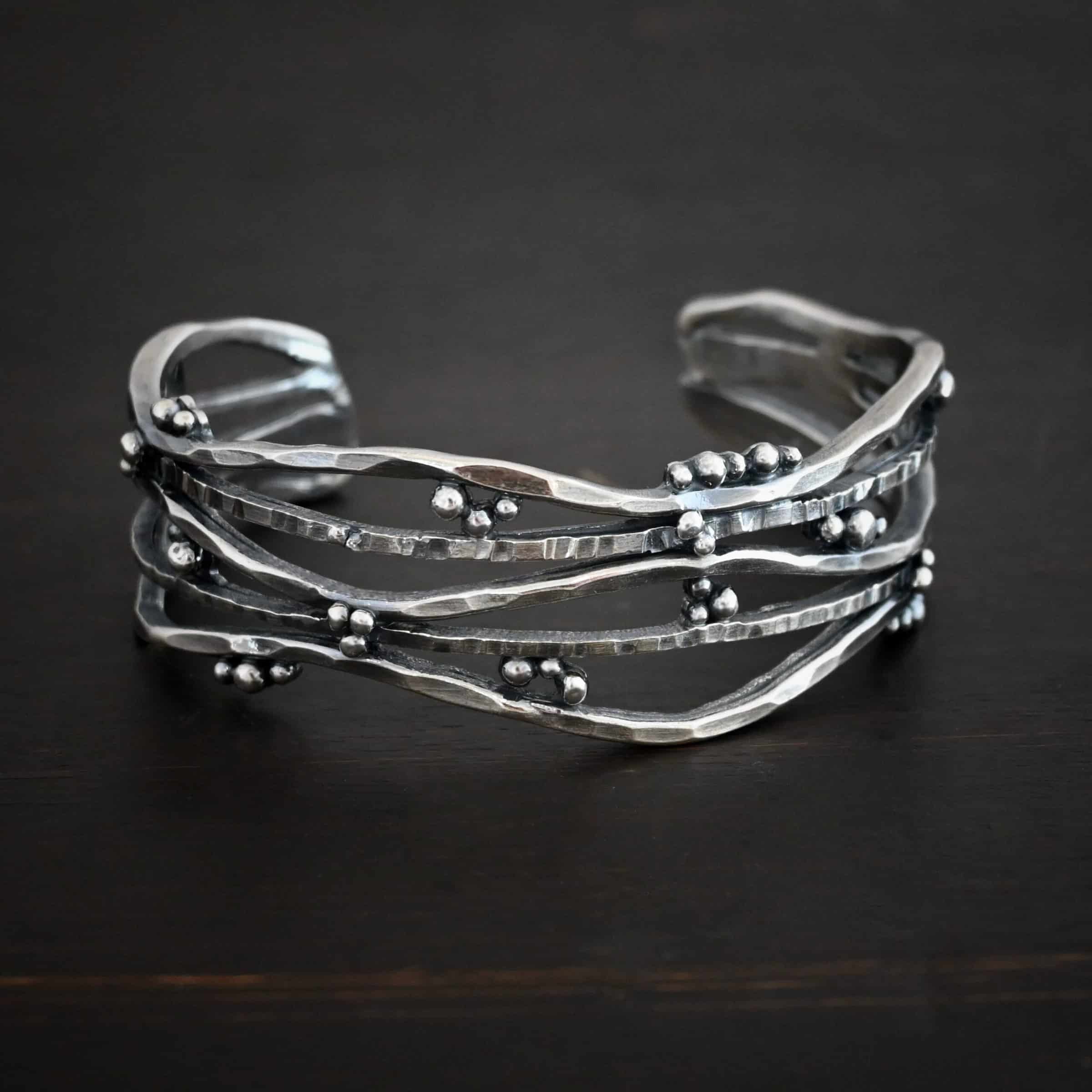 Wild Prairie Cuff Photo of the "Wild Prairie" Cuff. Made out of sterling silver. Cuff is made out of multiple thick, hammered lines of wavy silver. The silver connects at different points, throughout the cuff, but it all connects at the very back to create the cuff. There is silver ball detail around the entire cuff. Cuff is lying against a dark background.