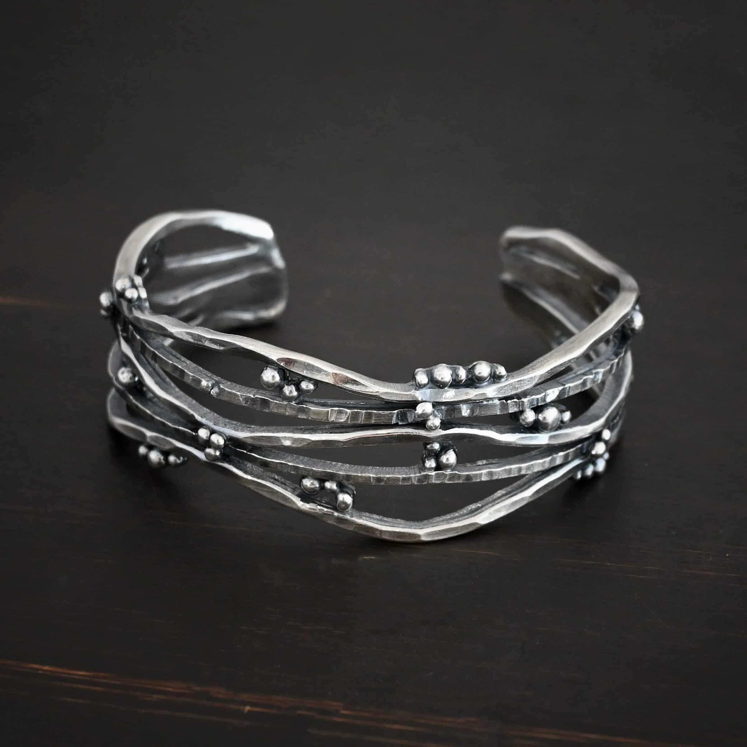 Wild Prairie Cuff Photo of the "Wild Prairie" Cuff. Made out of sterling silver. Cuff is made out of multiple thick, hammered lines of wavy silver. The silver connects at different points, throughout the cuff, but it all connects at the very back to create the cuff. There is silver ball detail around the entire cuff. Cuff is lying against a dark background.