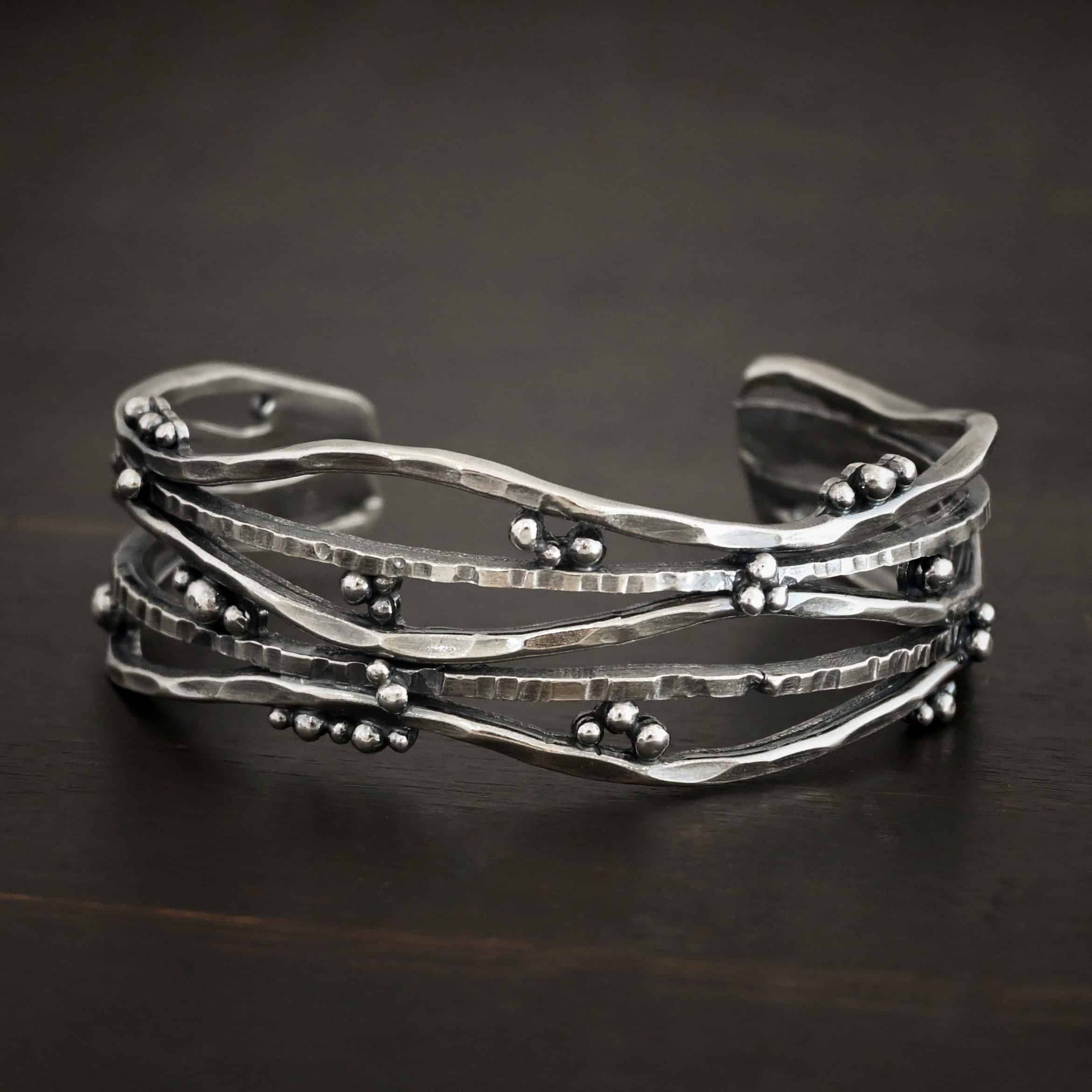 Wild Prairie Cuff Photo of the "Wild Prairie" Cuff. Made out of sterling silver. Cuff is made out of multiple thick, hammered lines of wavy silver. The silver connects at different points, throughout the cuff, but it all connects at the very back to create the cuff. There is silver ball detail around the entire cuff. Cuff is lying against a dark background.