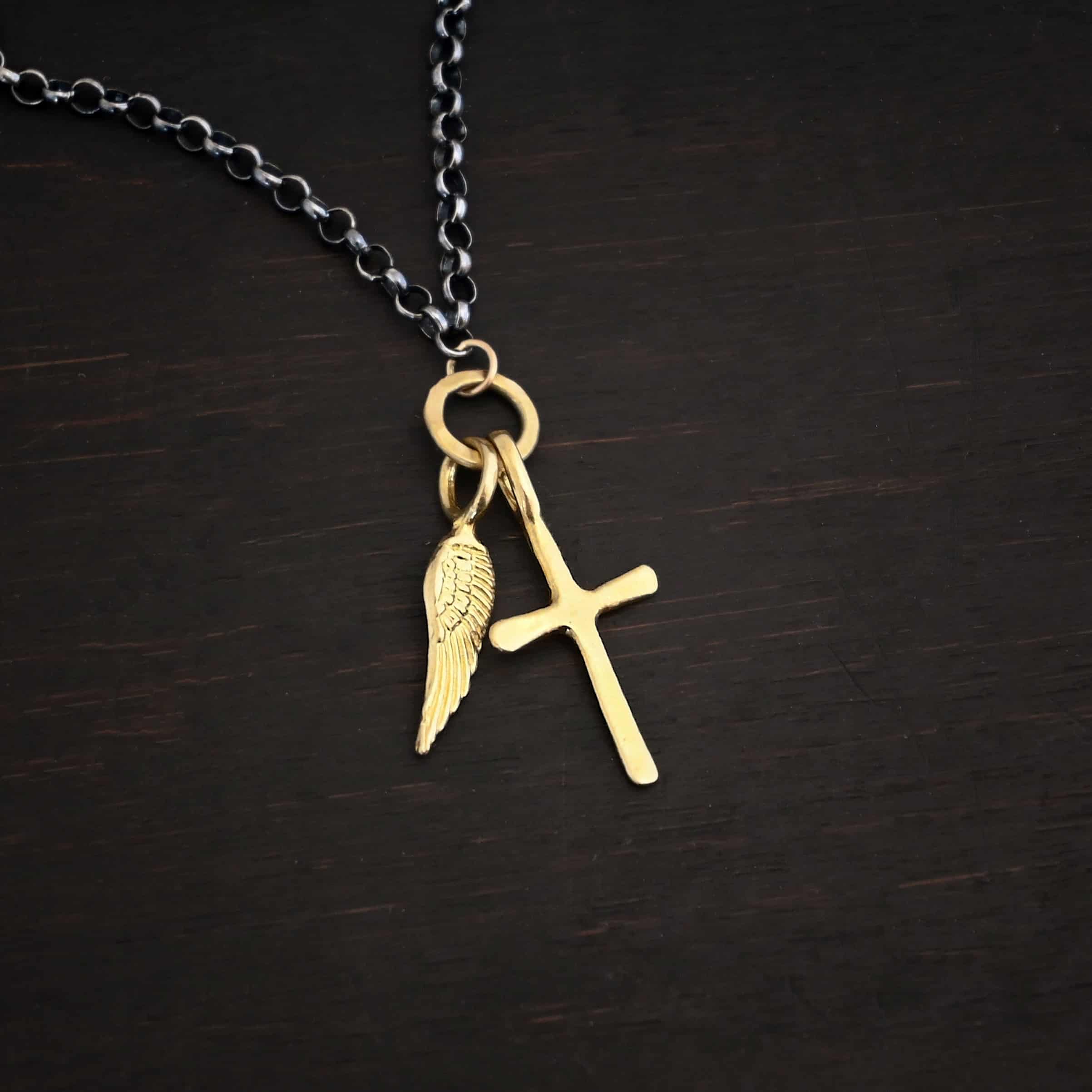 1 cross 1 wing ox 1 Photo of a cross and angel wing necklace. Made out of 18k gold. There is a small sized cross that is flat and smooth. It has a loop soldered to the top of it. There is a small sized angel wing pendant next to it, also with a soldered loop on the top. There is a chain that goes through the top of the necklace that is silver and oxidized. Necklace is lying against a dark background.