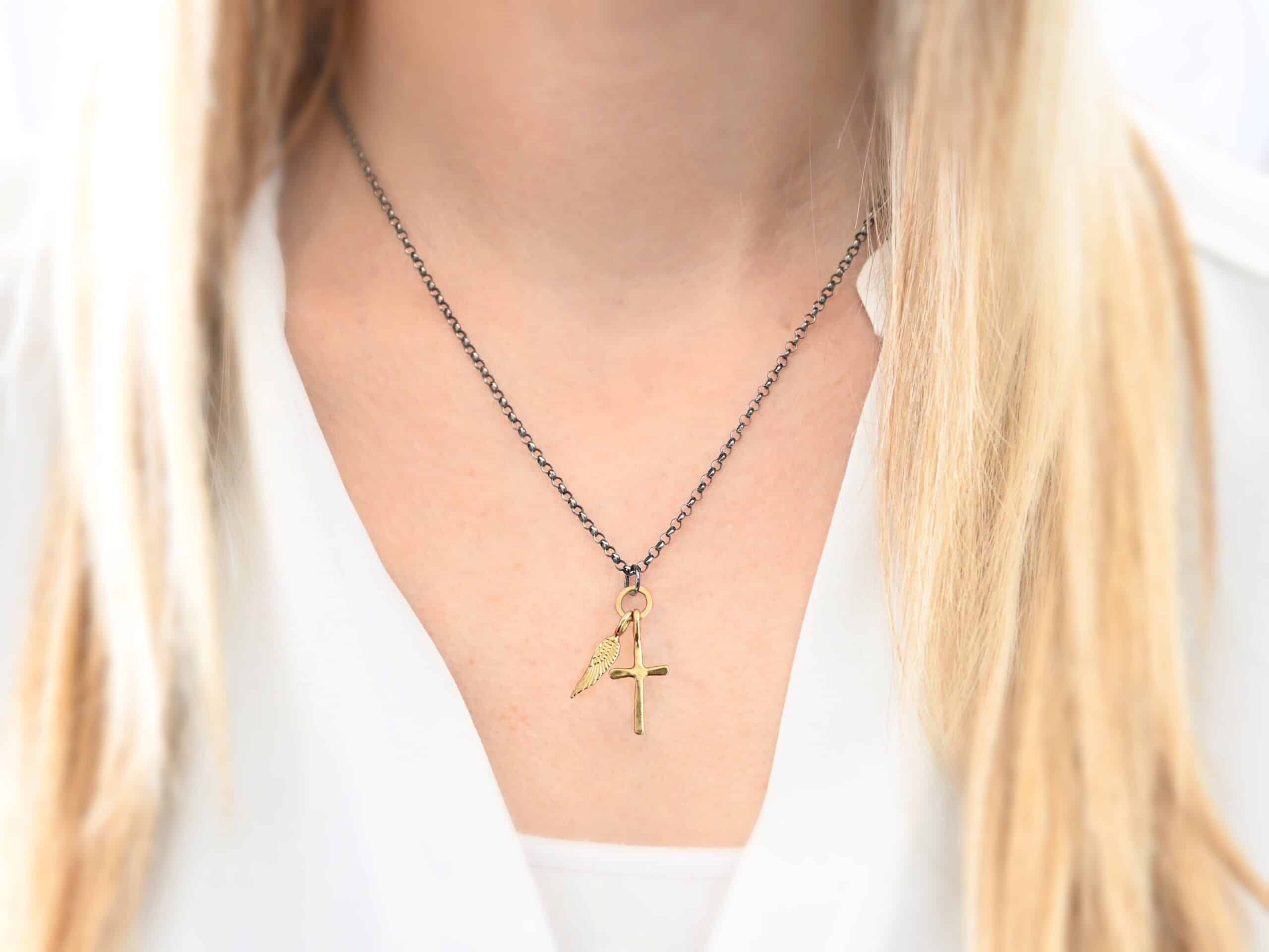 1 cross 1 wing ox 2 Photo of a cross and angel wing necklace. Made out of 18k gold. There is a small sized cross that is flat and smooth. It has a loop soldered to the top of it. There is a small sized angel wing pendant next to it, also with a soldered loop on the top. There is a chain that goes through the top of the necklace that is silver and oxidized. Necklace is worn by a model to demonstrate size.