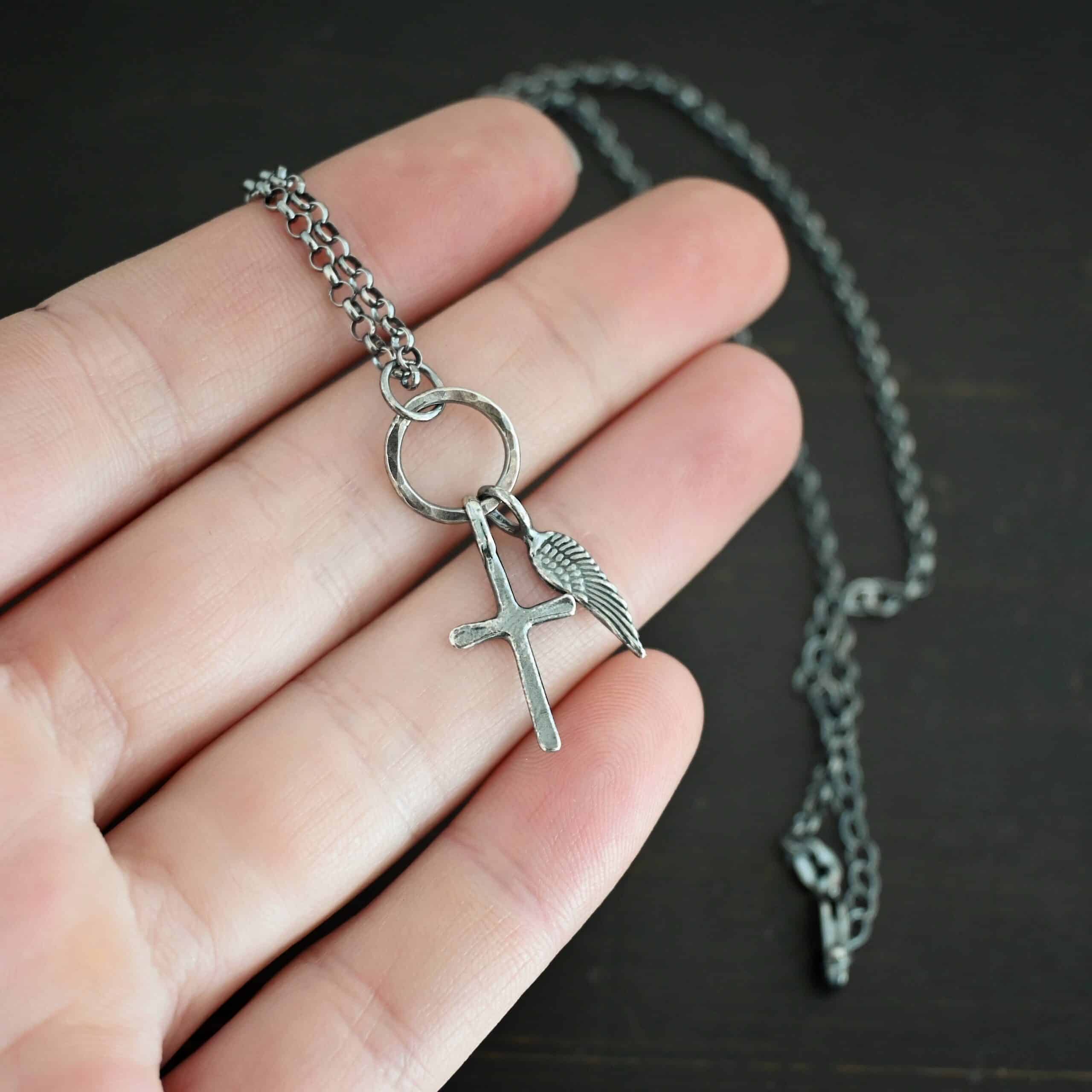 1 cross 1 wing ox 2 Photo of a cross and angel wing necklace. Made out of oxidized sterling silver. There is a small sized cross that is flat and smooth. It has a loop soldered to the top of it. There is a small sized angel wing pendant next to it, also with a soldered loop on the top. There is a chain that goes through the top of the necklace that is silver and oxidized. Necklace is lying against the fingertips of a hand.