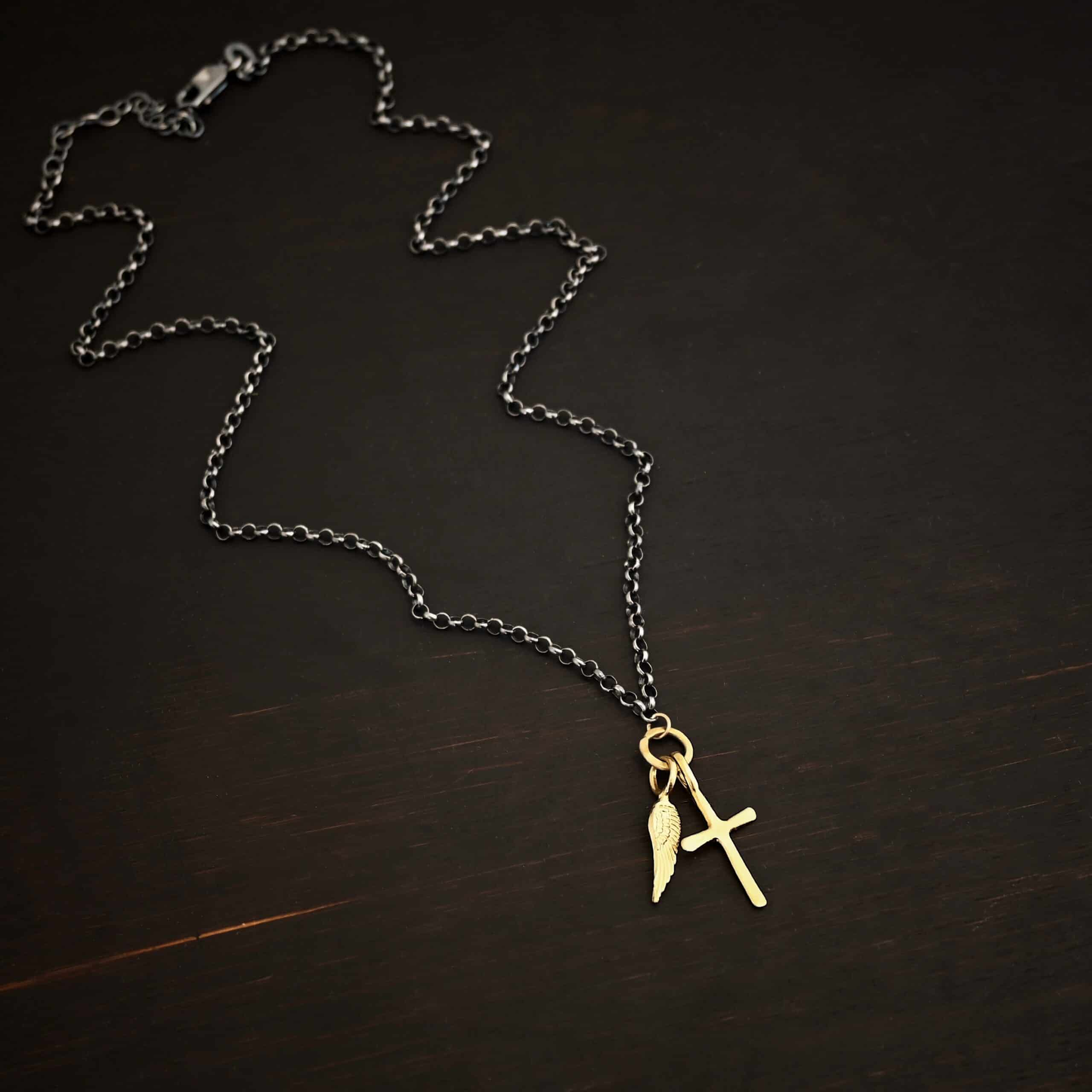 1 cross 1 wing ox 3 Photo of a cross and angel wing necklace. Made out of 18k gold. There is a small sized cross that is flat and smooth. It has a loop soldered to the top of it. There is a small sized angel wing pendant next to it, also with a soldered loop on the top. There is a chain that goes through the top of the necklace that is silver and oxidized. Necklace is lying against a dark background.
