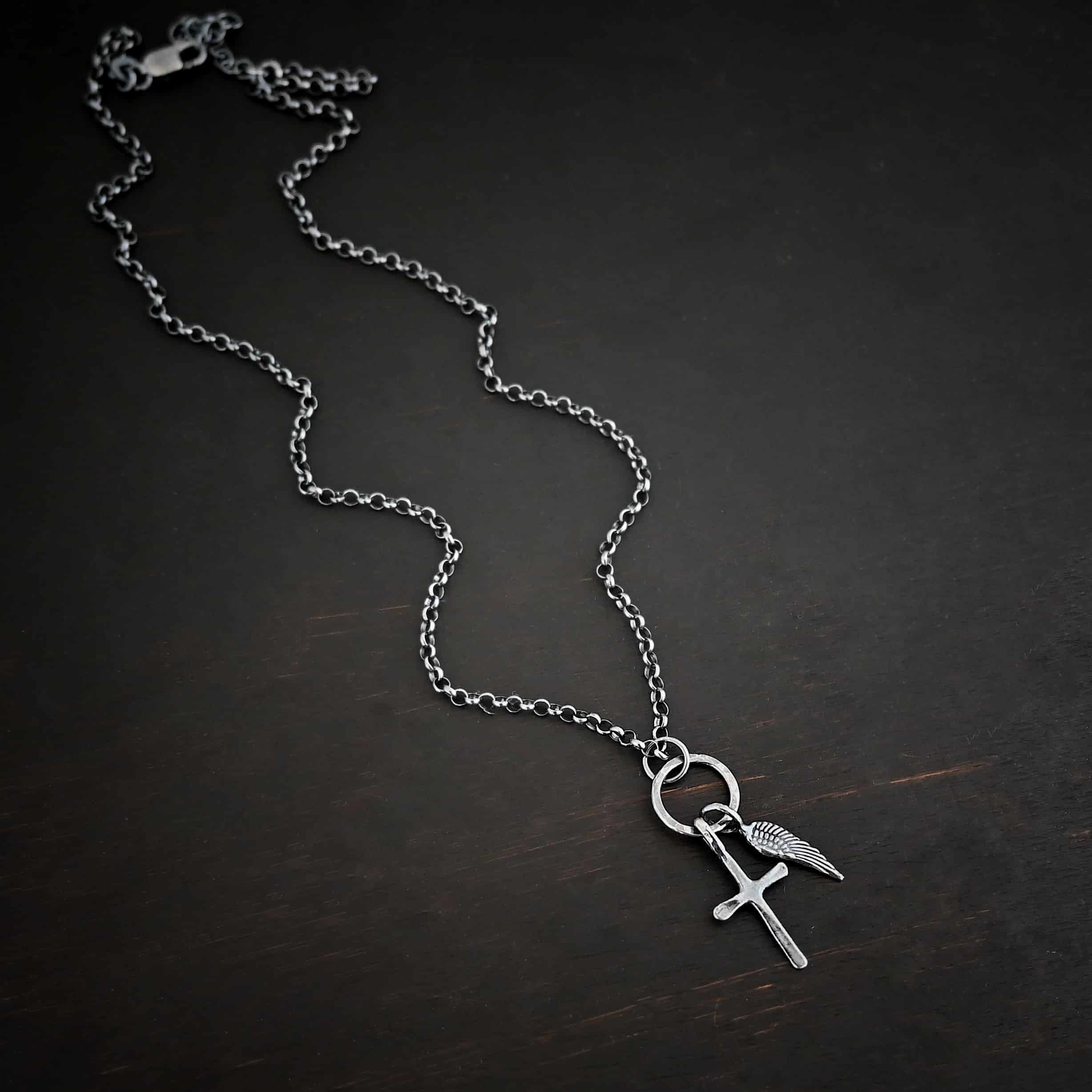 1 cross 1 wing ox 3 Photo of a cross and angel wing necklace. Made out of oxidized sterling silver. There is a small sized cross that is flat and smooth. It has a loop soldered to the top of it. There is a small sized angel wing pendant next to it, also with a soldered loop on the top. There is a chain that goes through the top of the necklace that is silver and oxidized. Necklace is lying against a dark background.