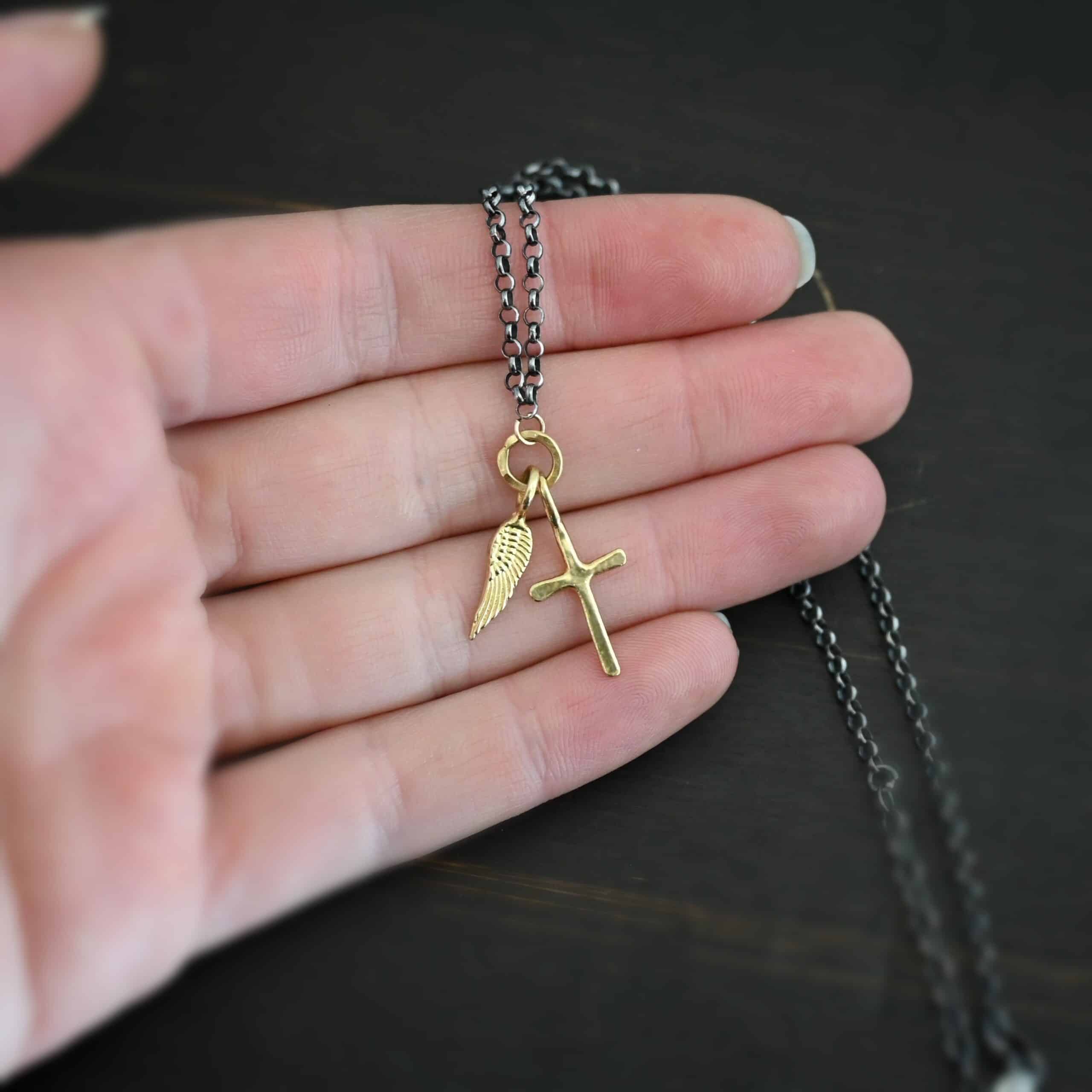 1 cross 1 wing ox 4 Photo of a cross and angel wing necklace. Made out of 18k gold. There is a small sized cross that is flat and smooth. It has a loop soldered to the top of it. There is a small sized angel wing pendant next to it, also with a soldered loop on the top. There is a chain that goes through the top of the necklace that is silver and oxidized. Necklace is lying against the fingertips of a hand.