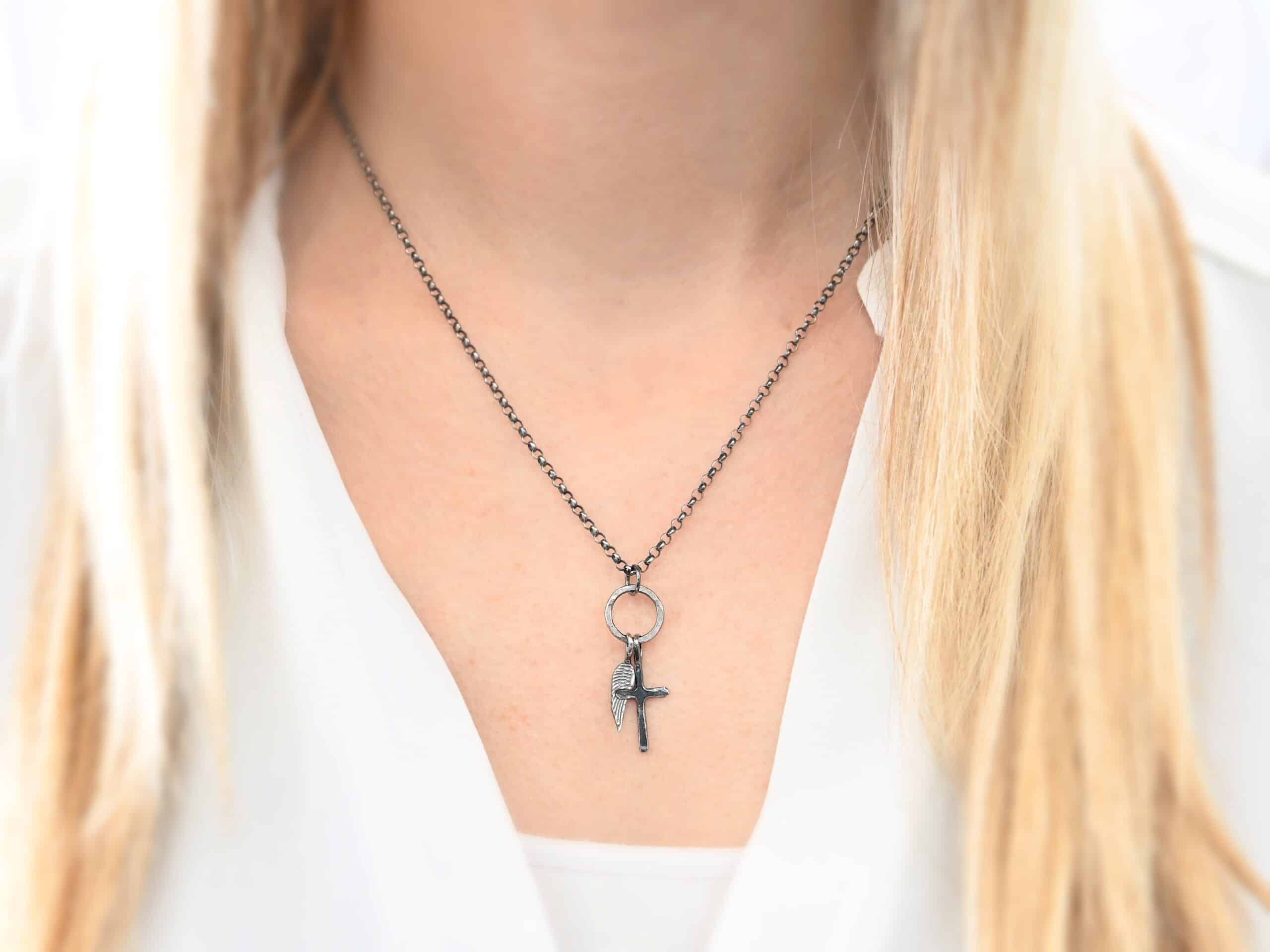 1 cross 1 wing ox Photo of a cross and angel wing necklace. Made out of oxidized sterling silver. There is a small sized cross that is flat and smooth. It has a loop soldered to the top of it. There is a small sized angel wing pendant next to it, also with a soldered loop on the top. There is a chain that goes through the top of the necklace that is silver and oxidized. Necklace is worn by a model.