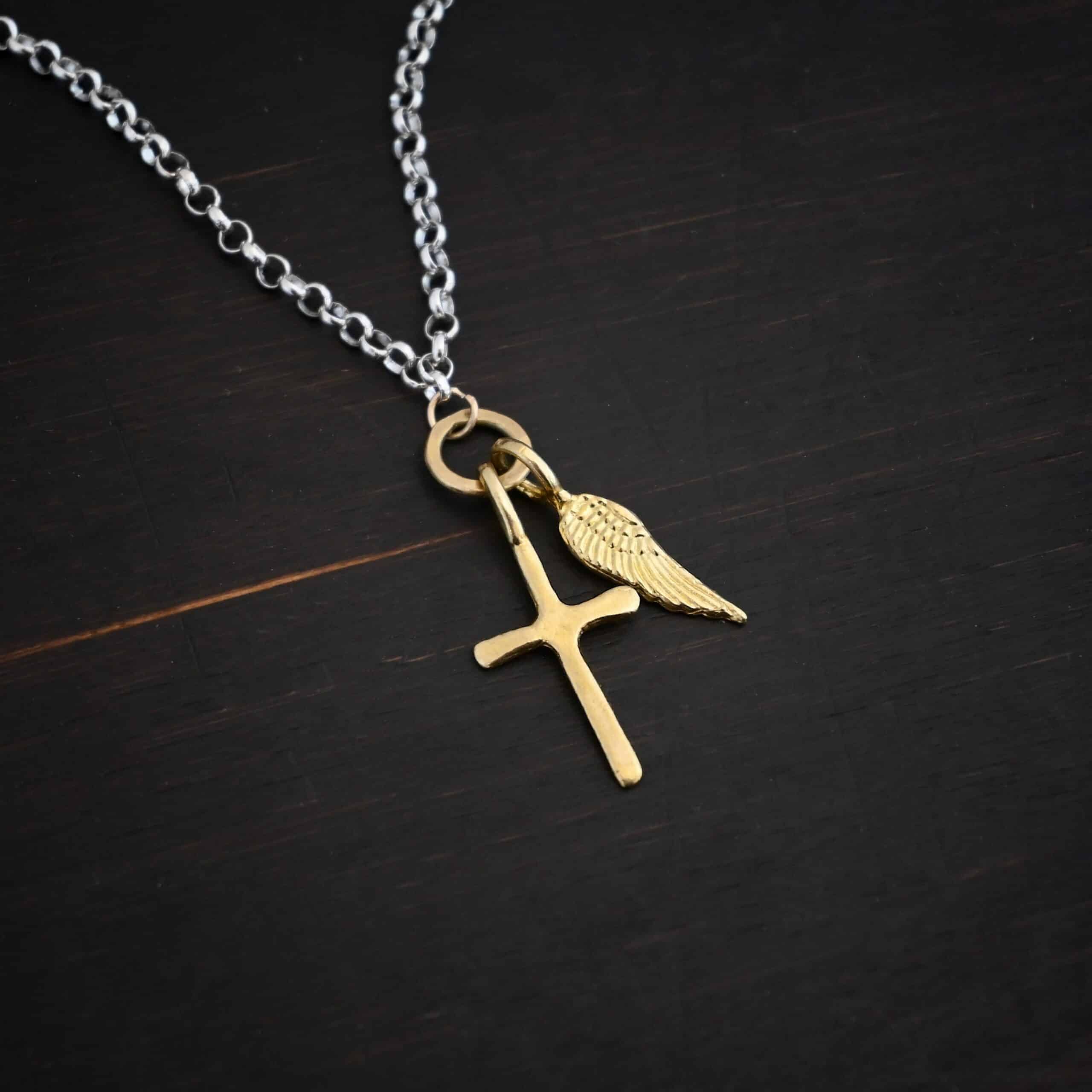 1 cross 1 wing shiny 1 Photo of a cross and angel wing necklace. Made out of 18k gold. There is a small sized cross that is flat and smooth. It has a loop soldered to the top of it. There is a small sized angel wing pendant next to it, also with a soldered loop on the top. There is a chain that goes through the top of the necklace that is silver and shiny. Necklace is lying against a dark background.