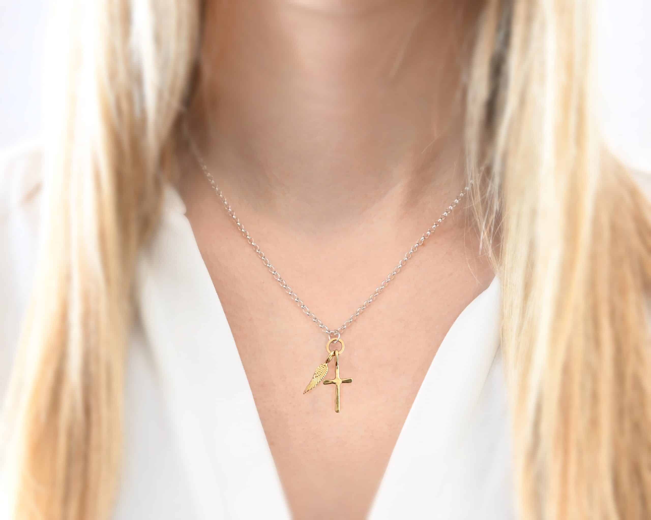 1 cross 1 wing shiny 2 Photo of a cross and angel wing necklace. Made out of 18k gold. There is a small sized cross that is flat and smooth. It has a loop soldered to the top of it. There is a small sized angel wing pendant next to it, also with a soldered loop on the top. There is a chain that goes through the top of the necklace that is silver and shiny. Necklace is worn by a model to demonstrate size.
