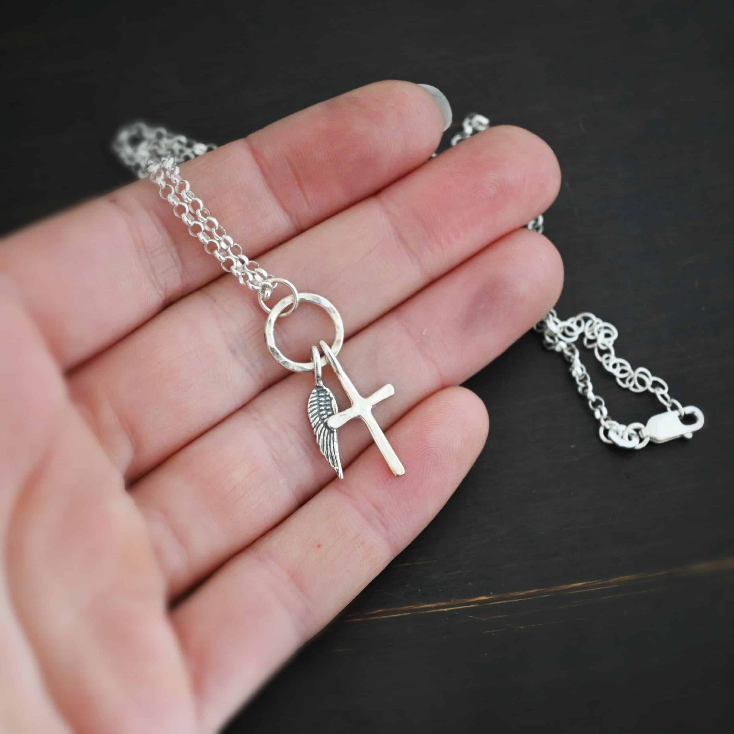 1 cross 1 wing shiny 2 Photo of a cross and angel wing necklace. Made out of shiny sterling silver. There is a small sized cross that is flat and smooth. It has a loop soldered to the top of it. There is a small sized angel wing pendant next to it, also with a soldered loop on the top. There is a chain that goes through the top of the necklace that is silver and shiny. Necklace is lying against the fingertips of a hand.