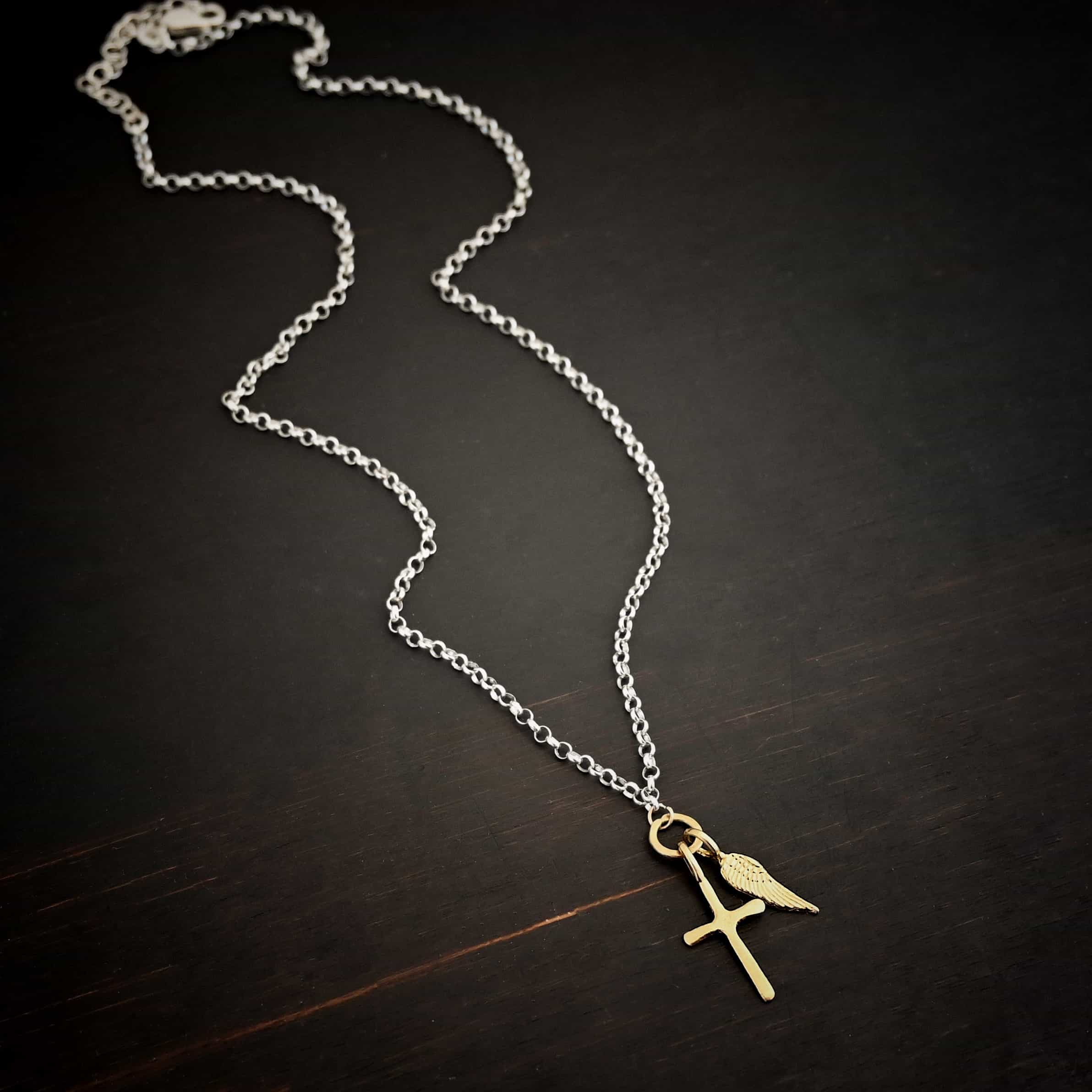 1 cross 1 wing shiny 3 Photo of a cross and angel wing necklace. Made out of 18k gold. There is a small sized cross that is flat and smooth. It has a loop soldered to the top of it. There is a small sized angel wing pendant next to it, also with a soldered loop on the top. There is a chain that goes through the top of the necklace that is silver and shiny. Necklace is lying against a dark background.