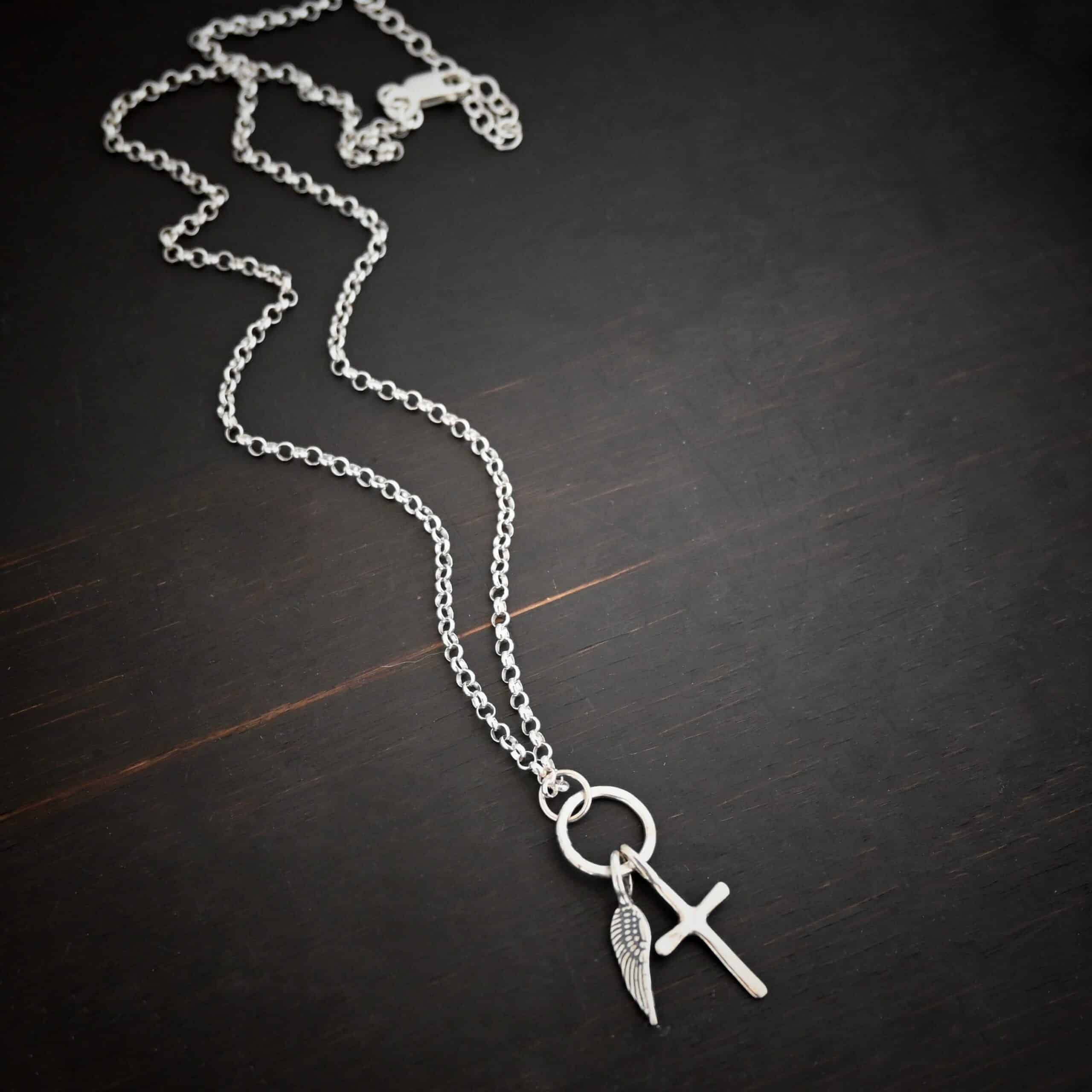 1 cross 1 wing shiny 3 Photo of a cross and angel wing necklace. Made out of shiny sterling silver. There is a small sized cross that is flat and smooth. It has a loop soldered to the top of it. There is a small sized angel wing pendant next to it, also with a soldered loop on the top. There is a chain that goes through the top of the necklace that is silver and shiny. Necklace is lying against a dark background.