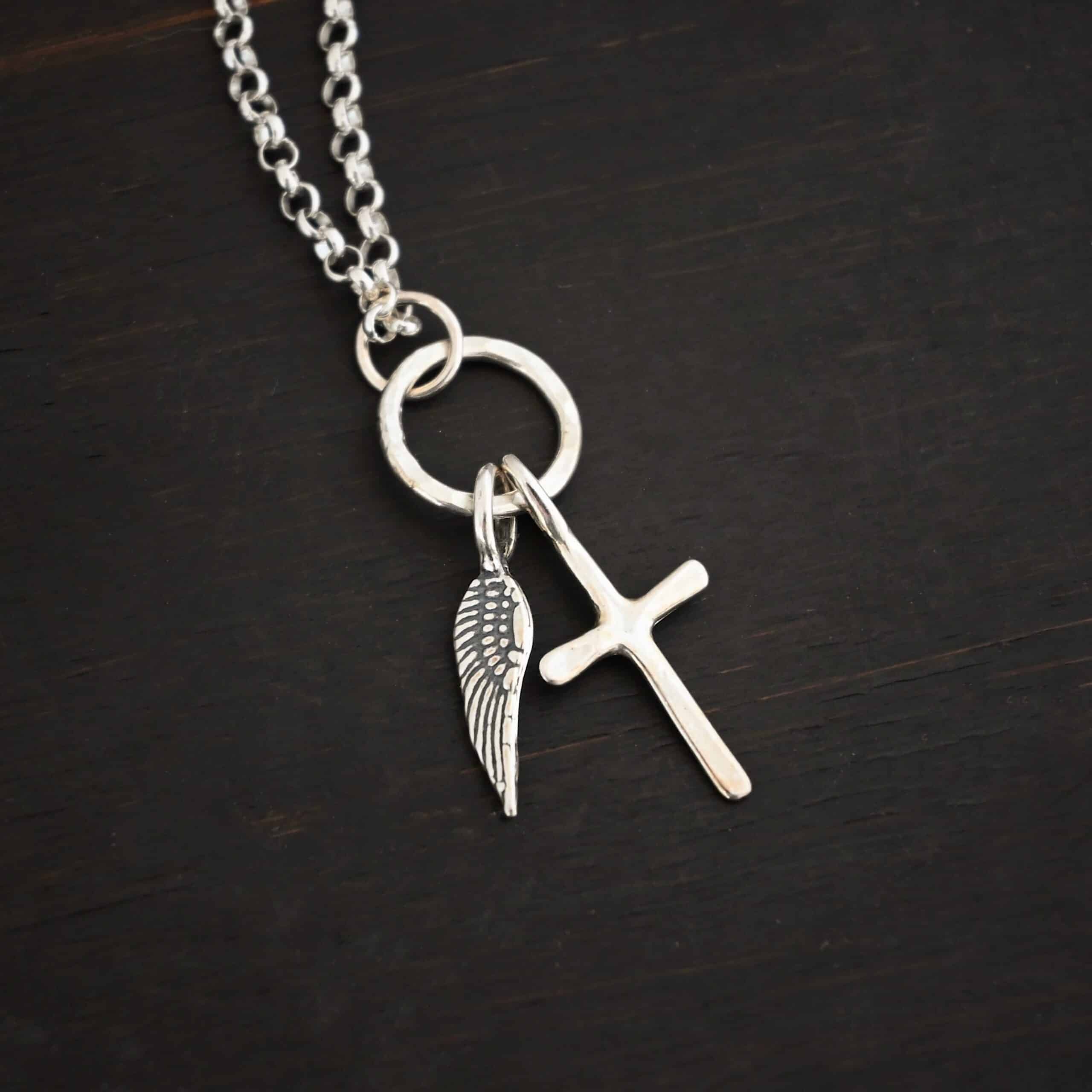 1 cross 1 wing shiny 4 Photo of a cross and angel wing necklace. Made out of shiny sterling silver. There is a small sized cross that is flat and smooth. It has a loop soldered to the top of it. There is a small sized angel wing pendant next to it, also with a soldered loop on the top. There is a chain that goes through the top of the necklace that is silver and shiny. Necklace is lying against a dark background.
