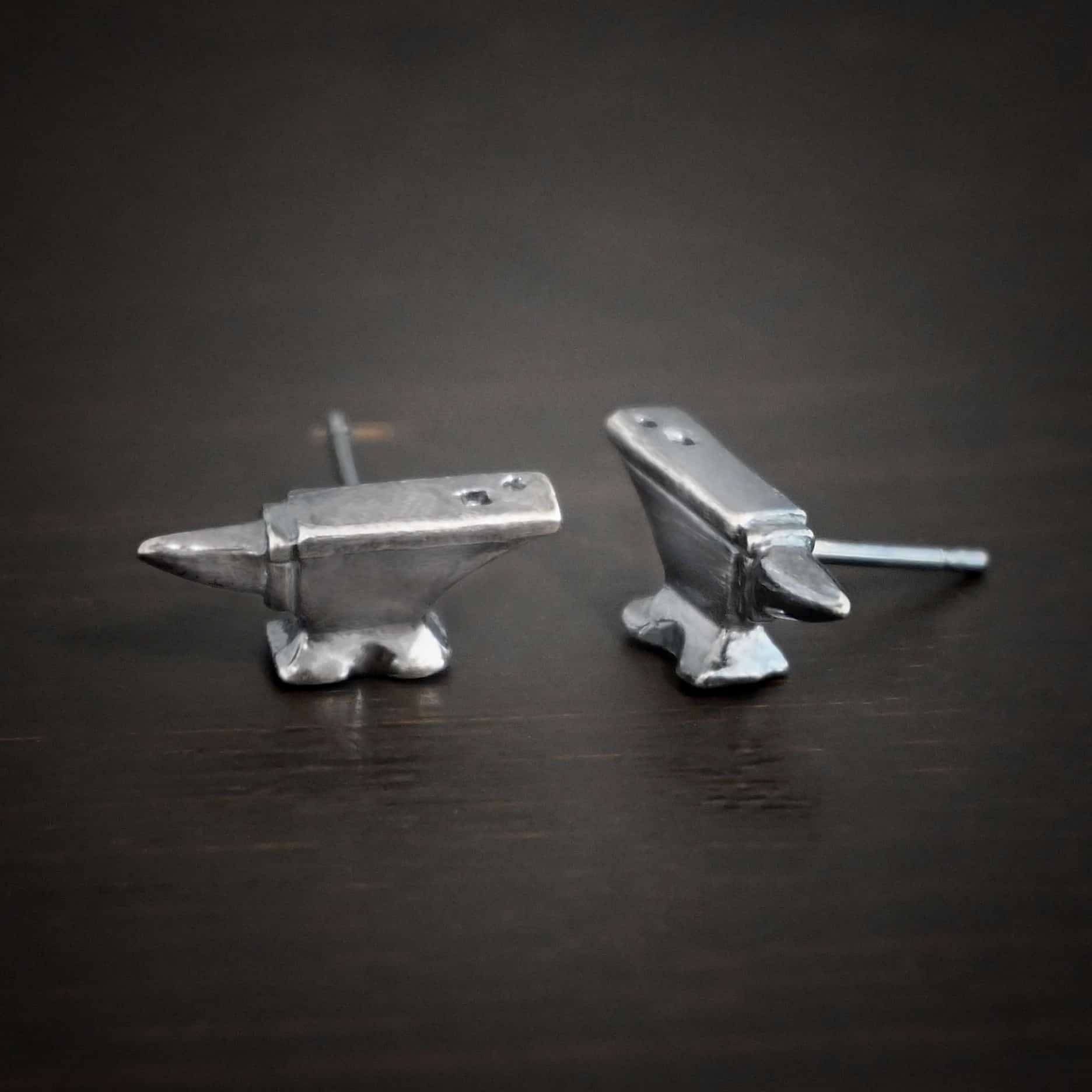 Photo of the "Silversmith" Earrings. Earrings are made out of sterling silver and have post ear wires. Earrings have small anvil charms that the post ear wires are attached to. Earrings are lying against a dark background.