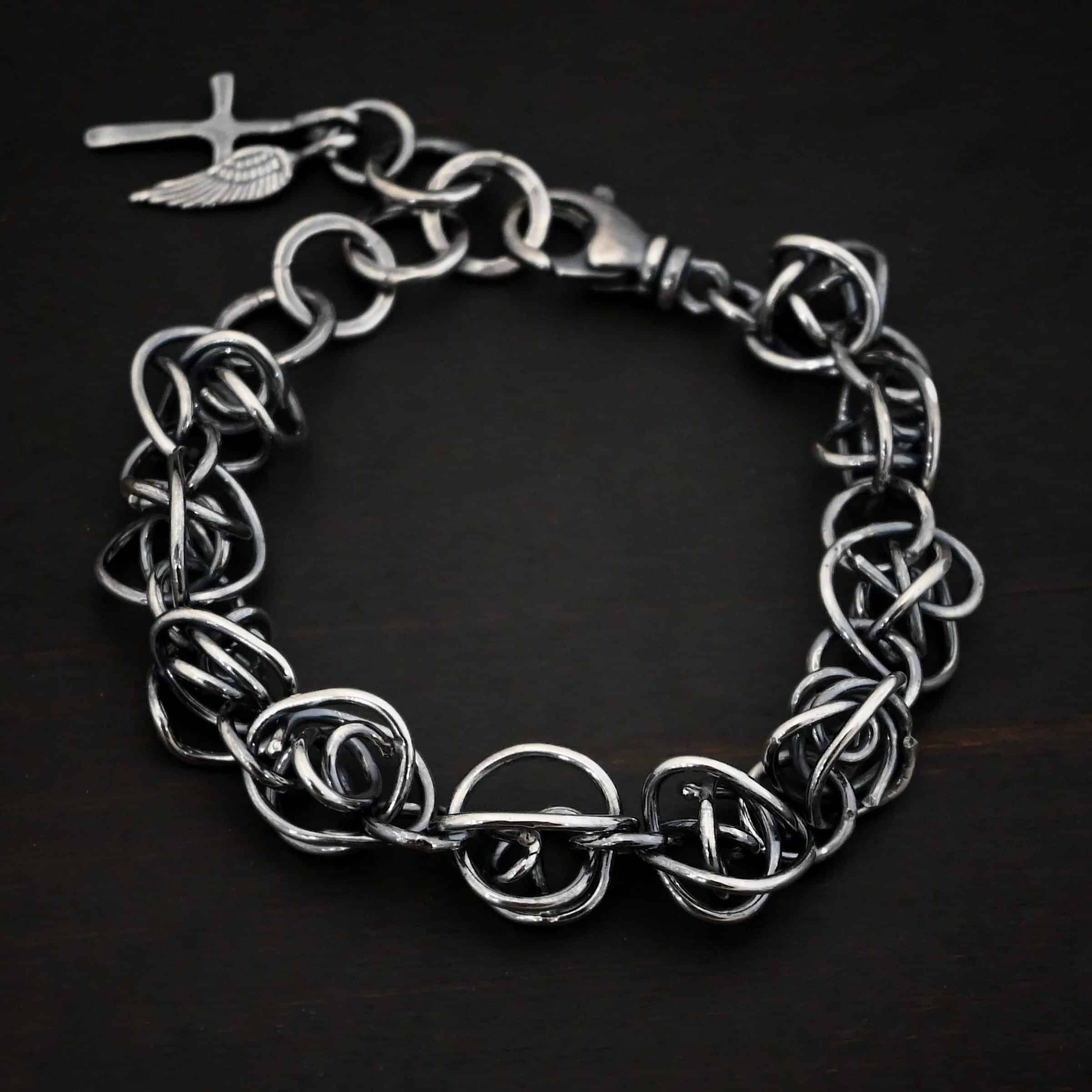 Photo of the "Basket Case" Bracelet. Made out of sterling silver. There are ten, medium sized balls of silver that are all connected closely by jump rings. Each ball is a jumble of sterling silver that wraps around itself multiple times, so it looks crazy and tangled. All of the balls are so close together, that it looks like a continuous wrap. Bracelet has a lobster claw clasp, and a cross and angel wing charm on the back. Bracelet is resting against a dark background.