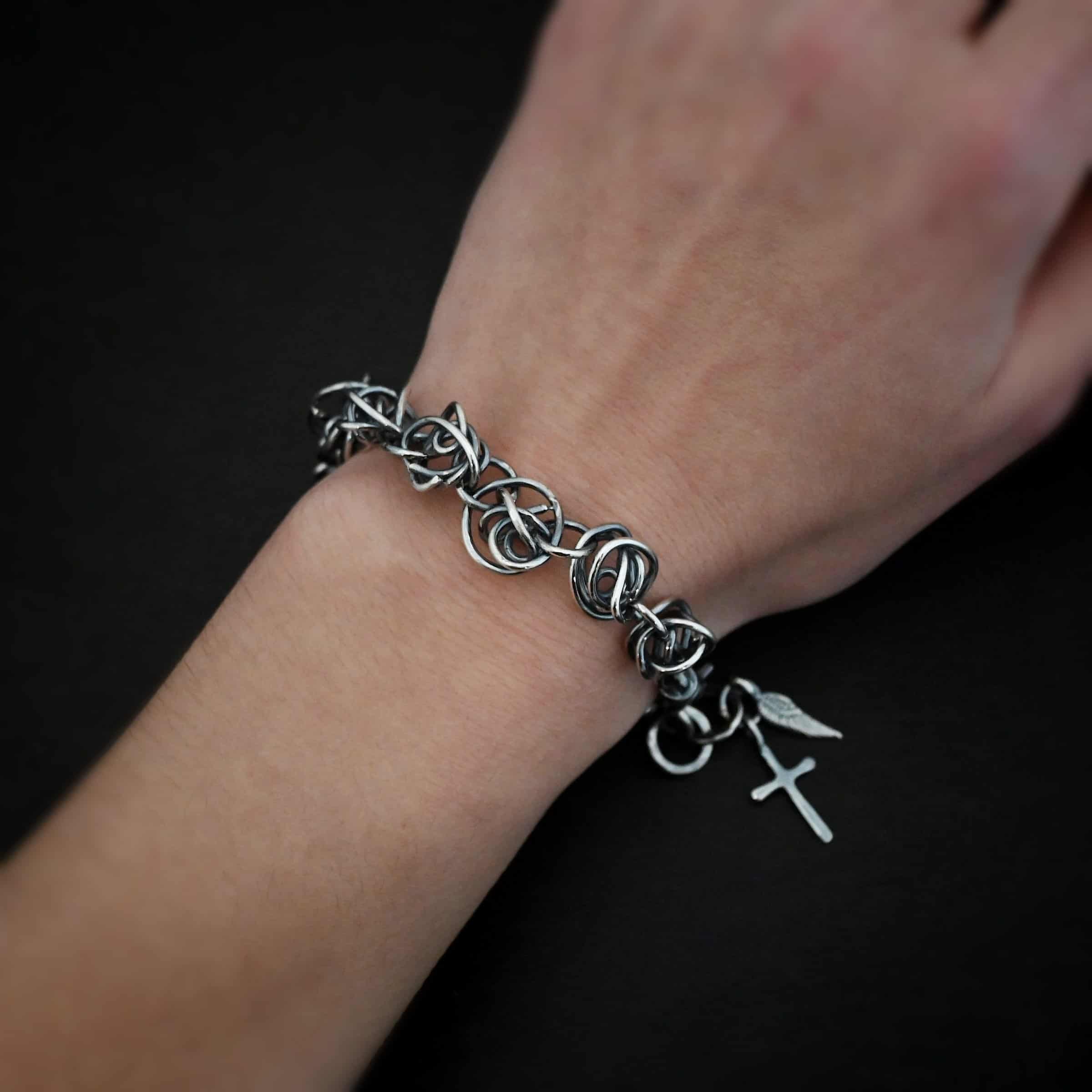 Photo of the "Basket Case" Bracelet. Made out of sterling silver. There are ten, medium sized balls of silver that are all connected closely by jump rings. Each ball is a jumble of sterling silver that wraps around itself multiple times, so it looks crazy and tangled. All of the balls are so close together, that it looks like a continuous wrap. Bracelet has a lobster claw clasp, and a cross and angel wing charm on the back. Bracelet is worn by a model up close on their wrist.