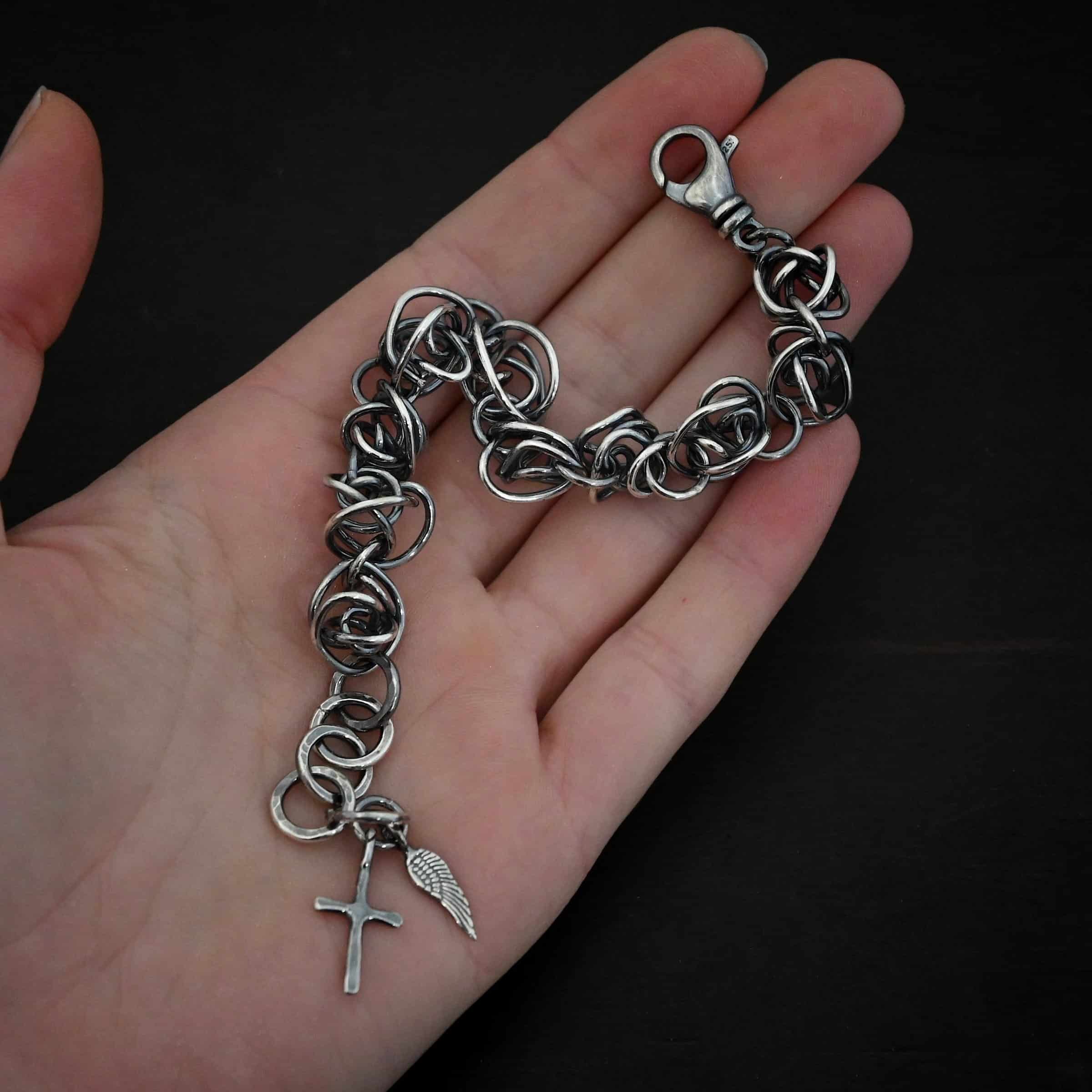 Photo of the "Basket Case" Bracelet. Made out of sterling silver. There are ten, medium sized balls of silver that are all connected closely by jump rings. Each ball is a jumble of sterling silver that wraps around itself multiple times, so it looks crazy and tangled. All of the balls are so close together, that it looks like a continuous wrap. Bracelet has a lobster claw clasp, and a cross and angel wing charm on the back. Bracelet is lying on the palm and fingertips of a hand.