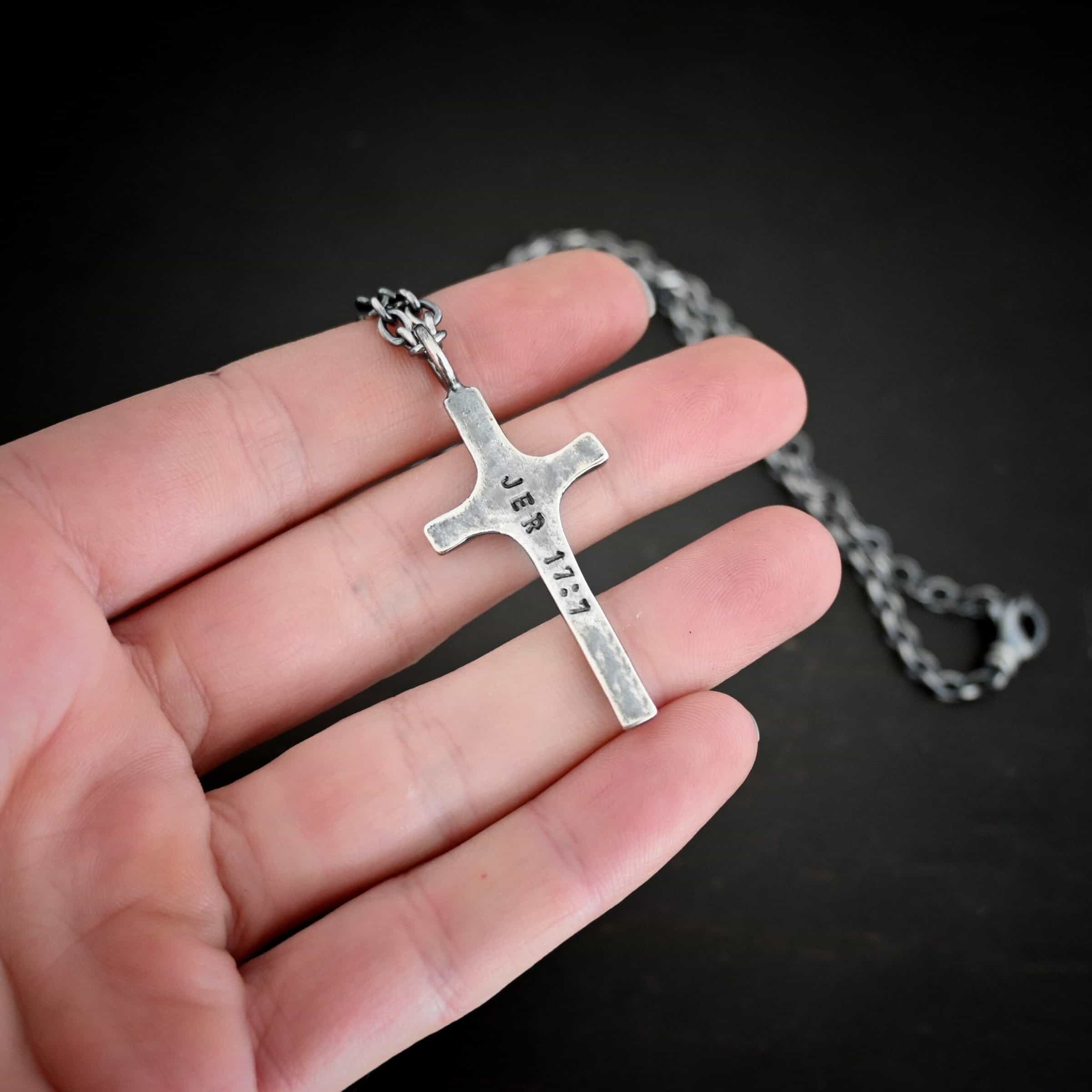 Photo of the back of a custom necklace. Made out of sterling silver. There is a medium sized cross that is made out of smooth, thick sterling silver. It is oxidized and brushed lightly and has a loop soldered to the top of it. The words "Jer 17:7" stamped into it. There is a chain that goes through the top of the necklace. Necklace is lying against the fingertips of a hand.