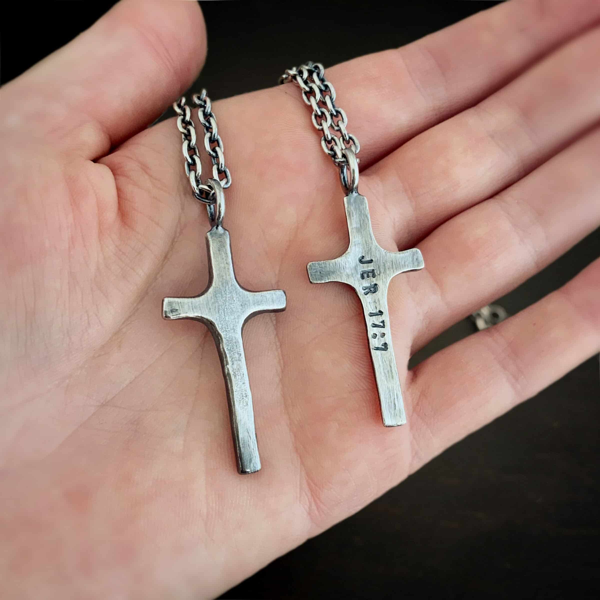 Photo of the back of the"King Of Kings" Necklace. Made out of sterling silver. There is a medium sized cross that is made out of smooth, thick sterling silver. It is oxidized and brushed lightly and has a loop soldered to the top of it. The words "Jer 17:7" stamped into it. There is a chain that goes through the top of the necklace. Necklace is lying against the palm of a hand, along with the front of a similar necklace.