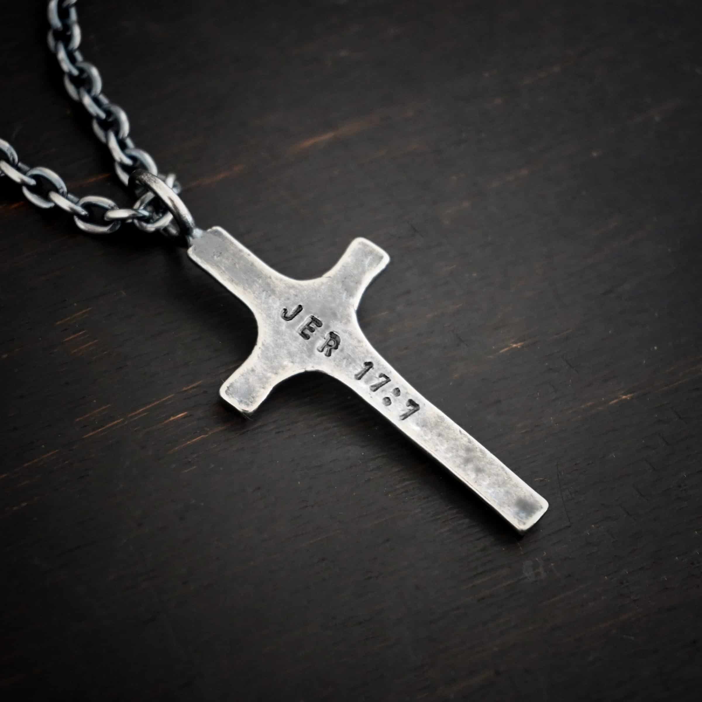 Photo of the back of a custom necklace. Made out of sterling silver. There is a medium sized cross that is made out of smooth, thick sterling silver. It is oxidized and brushed lightly and has a loop soldered to the top of it. The words "Jer 17:7" stamped into it. There is a chain that goes through the top of the necklace. Necklace is lying against a dark background.