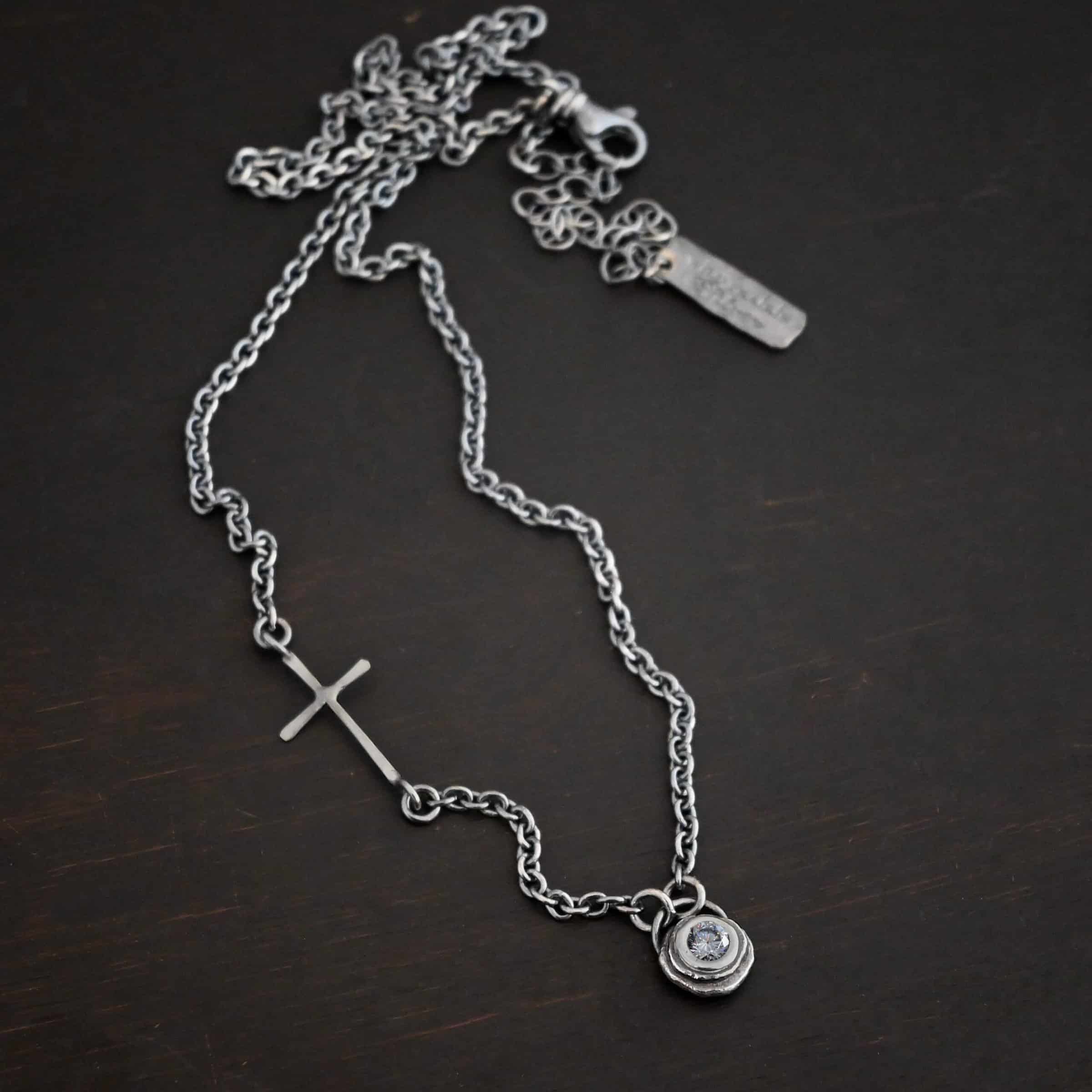 Devotion Necklace Silver Photo of the "Devotion" Necklace. There is a small diamond in the center of a small circle pendant. The diamond is set in silver. There are two layers of sterling silver that are both hammered flat dots. One layer of silver is smaller, and sits on top of the bigger layer of silver. The silver is on the very top of the pendant. There is a small loop soldered to the back of the necklace in which two silver jump rings hang from. Each end of the chain hangs on the jump rings. There is a large silver cross that is attached on the left side of the chain that is attached to the chain with two jump rings. The end and top of the cross have a loop. The bottom loop attaches to a jump ring on the chain, as well as the top loop, so the cross is fully connected to the chain. Necklace is lying against a dark background.