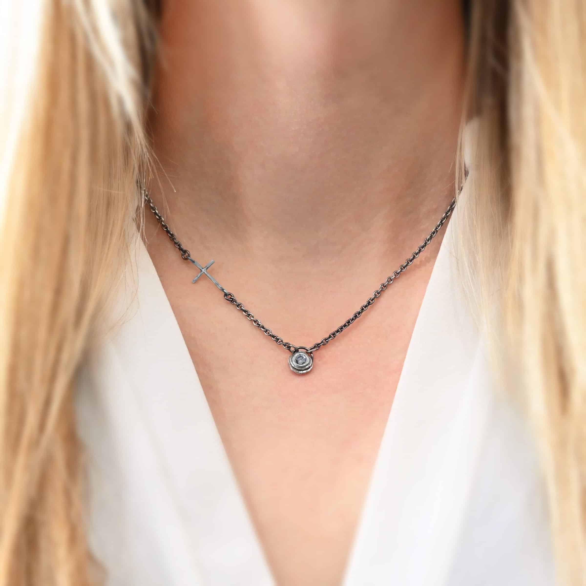 Devotion Necklace Silver Photo of the "Devotion" Necklace. There is a small diamond in the center of a small circle pendant. The diamond is set in silver. There are two layers of sterling silver that are both hammered flat dots. One layer of silver is smaller, and sits on top of the bigger layer of silver. The silver is on the very top of the pendant. There is a small loop soldered to the back of the necklace in which two silver jump rings hang from. Each end of the chain hangs on the jump rings. There is a large silver cross that is attached on the left side of the chain that is attached to the chain with two jump rings. The end and top of the cross have a loop. The bottom loop attaches to a jump ring on the chain, as well as the top loop, so the cross is fully connected to the chain. Necklace is worn by a model to demonstrate size.