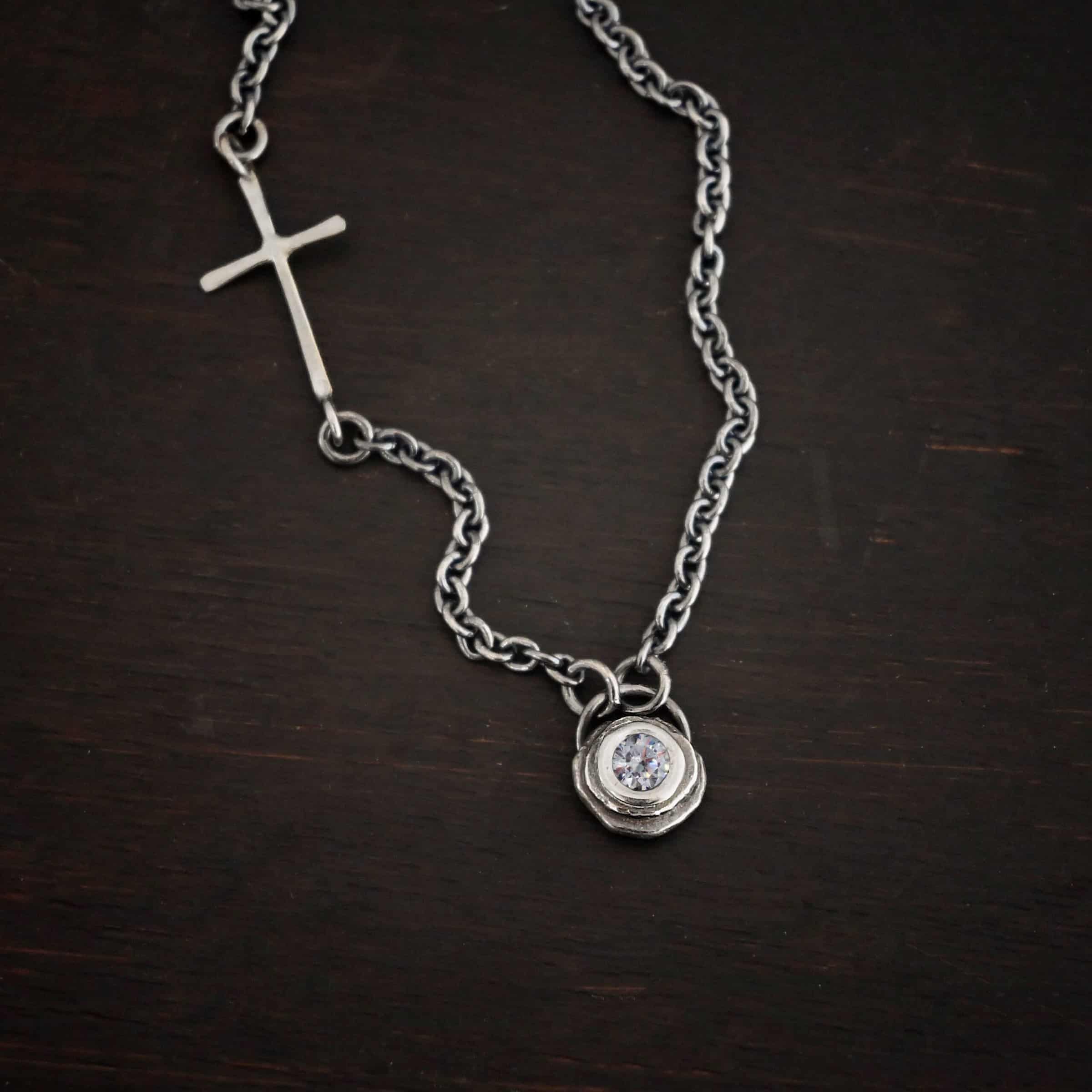 Devotion Necklace Silver Photo of the "Devotion" Necklace. There is a small diamond in the center of a small circle pendant. The diamond is set in silver. There are two layers of sterling silver that are both hammered flat dots. One layer of silver is smaller, and sits on top of the bigger layer of silver. The silver is on the very top of the pendant. There is a small loop soldered to the back of the necklace in which two silver jump rings hang from. Each end of the chain hangs on the jump rings. There is a large silver cross that is attached on the left side of the chain that is attached to the chain with two jump rings. The end and top of the cross have a loop. The bottom loop attaches to a jump ring on the chain, as well as the top loop, so the cross is fully connected to the chain. Necklace is lying against a dark background.