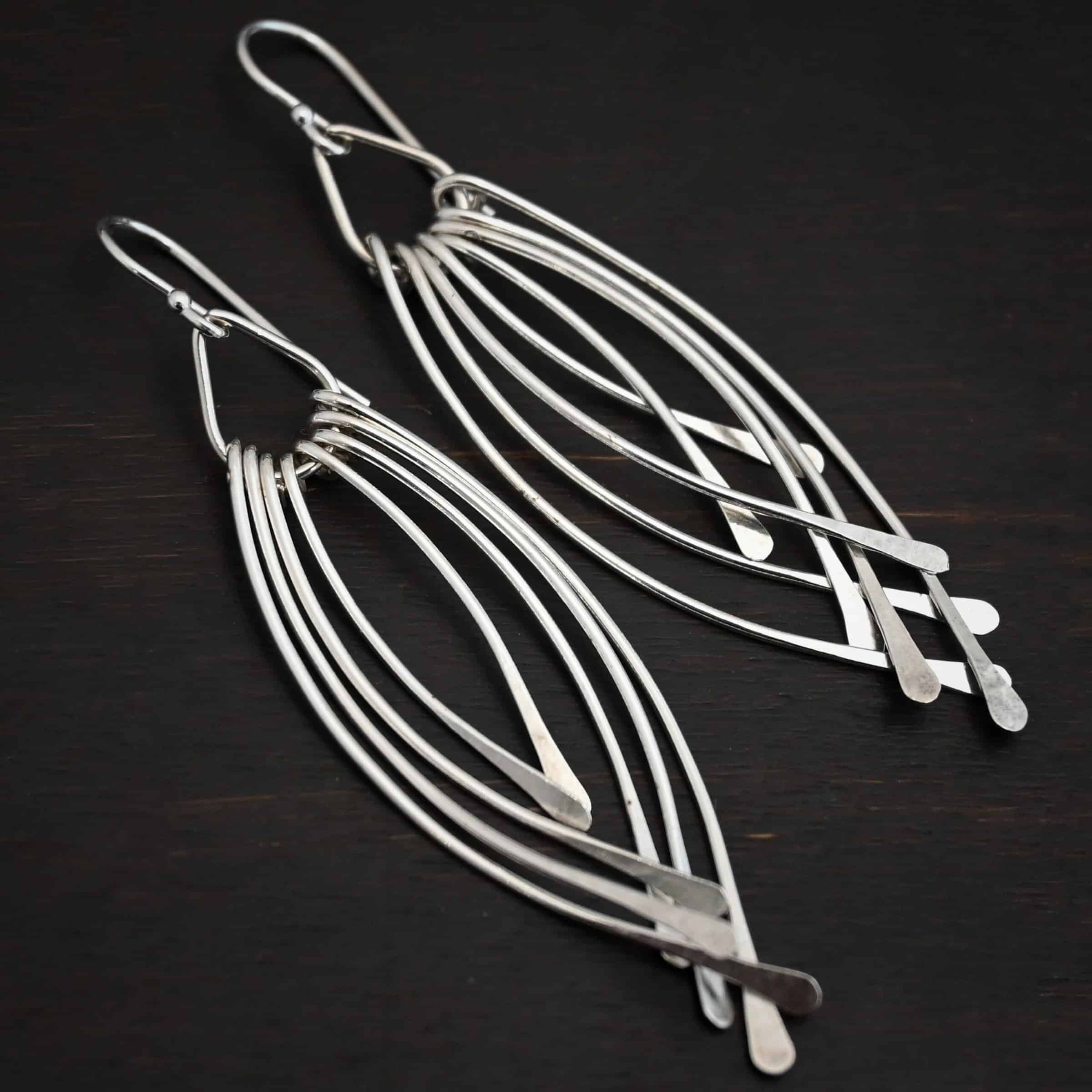 Prairie Wind Earrings Photo of the "Prairie Wind" Earrings. Made out of sterling silver and oxidized. Earrings have french hook ear wires. Attached to the bottom of the ear wires is a teardrop shaped loop of silver. Attached to the bottom of the teardrop loop are eight pieces of silver that all have hammered paddle ends. Four pieces of silver curve towards the right, and the other four pieces curve towards the left, so they overlap each other. Earrings are lying against a dark background.