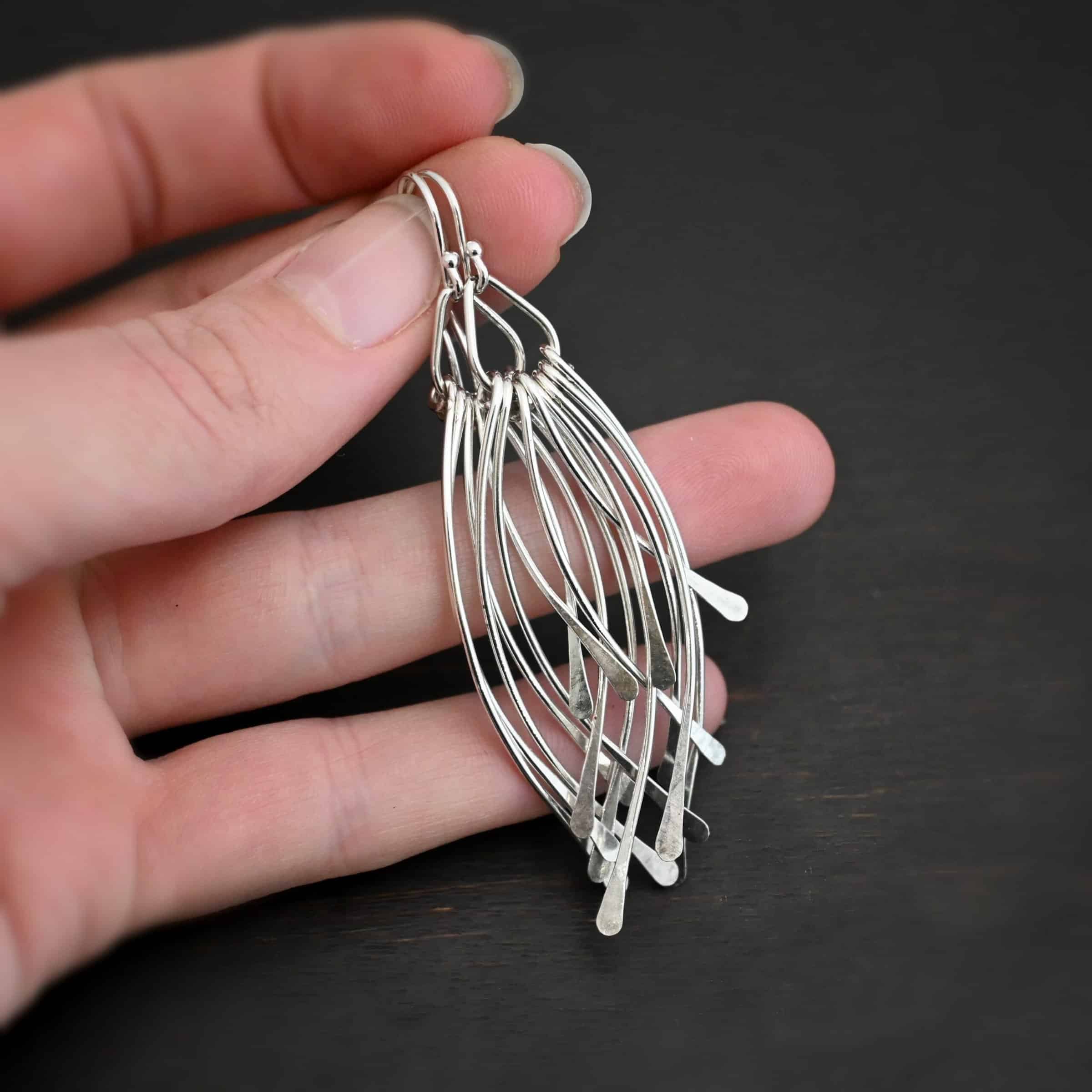 Prairie Wind Earrings Photo of the "Prairie Wind" Earrings. Made out of sterling silver and oxidized. Earrings have french hook ear wires. Attached to the bottom of the ear wires is a teardrop shaped loop of silver. Attached to the bottom of the teardrop loop are eight pieces of silver that all have hammered paddle ends. Four pieces of silver curve towards the right, and the other four pieces curve towards the left, so they overlap each other. Earrings are held in the fingertips of a hand.