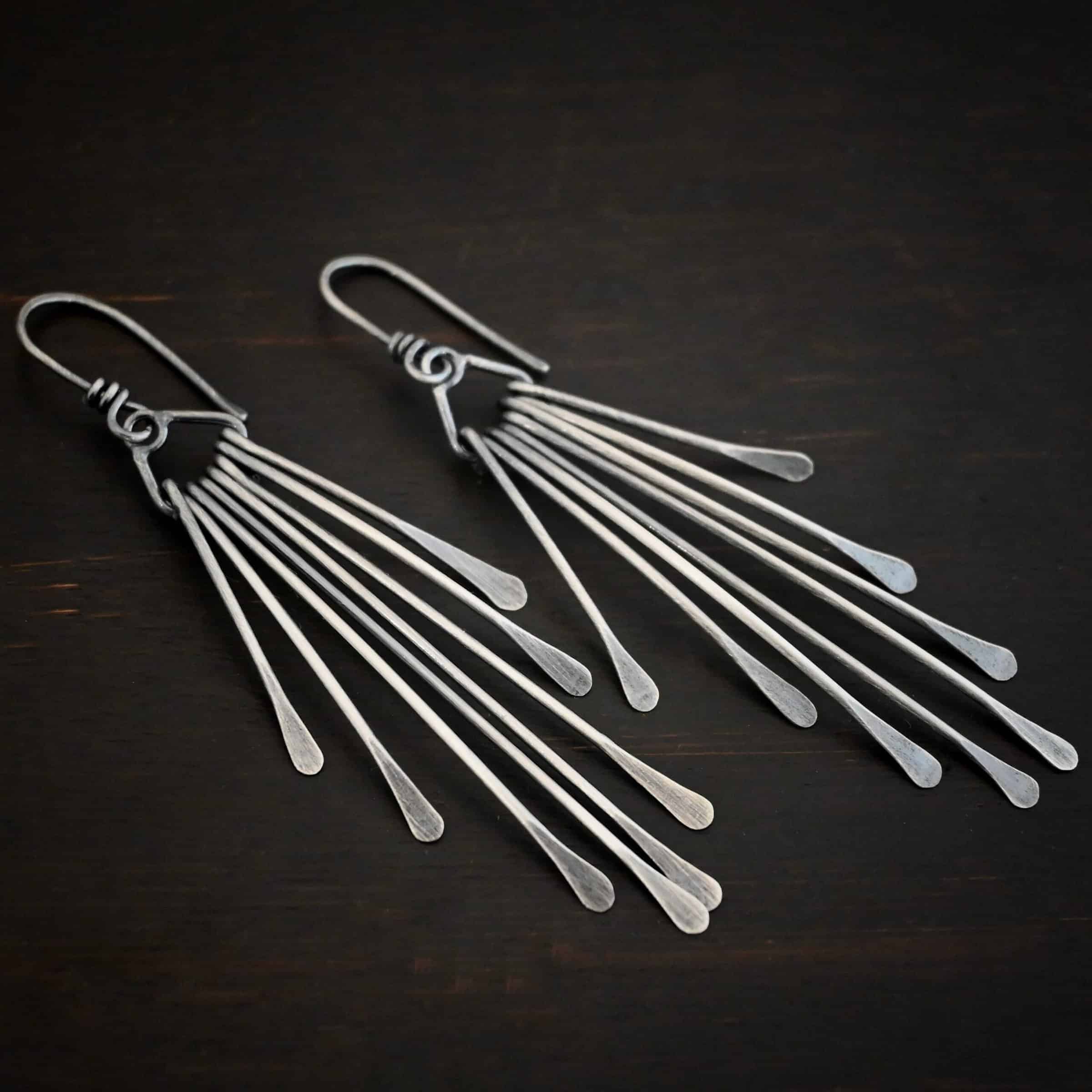 Photo of the "Marilyn Monroe" Earrings. Made out of sterling silver. Earrings have french hook ear wires. Attached to the bottom loop of the ear wires is a teardrop shaped piece of silver. Hanging from this piece are eight separate pieces of silver, all with hammered paddle ends. They all hang straight down. The shortest ones are on the outside, and they get progressively longer until the middle. So they go from short, to long, and back to short. Earrings are lying against a dark background.