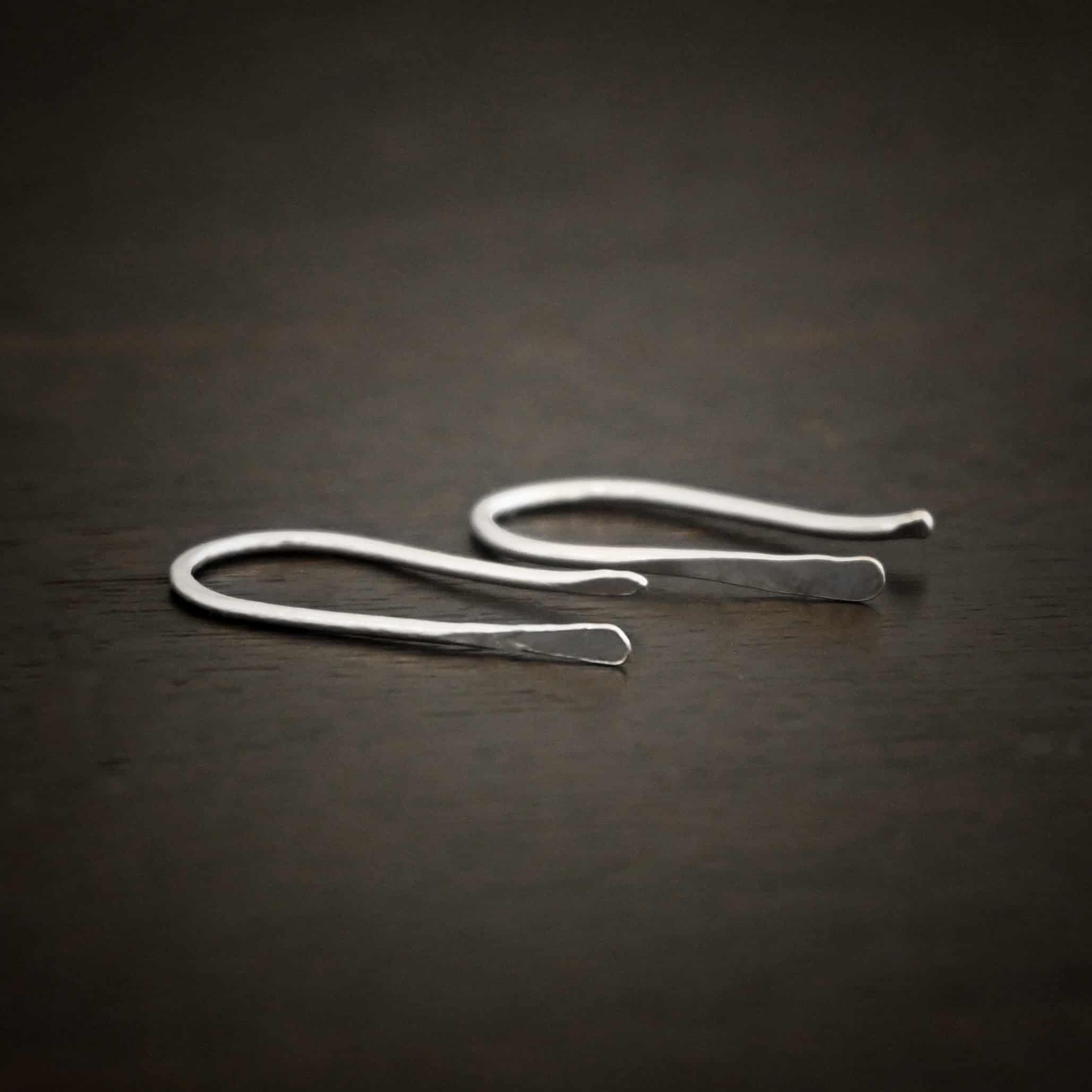 Roadies Photo of the "Roadie" Earrings. Made out of sterling silver. Earrings are very simple. They are an ear wire with one end lightly hammered and tipped and the other end of the earring is a hammered paddle. Earrings are lying against a dark background.