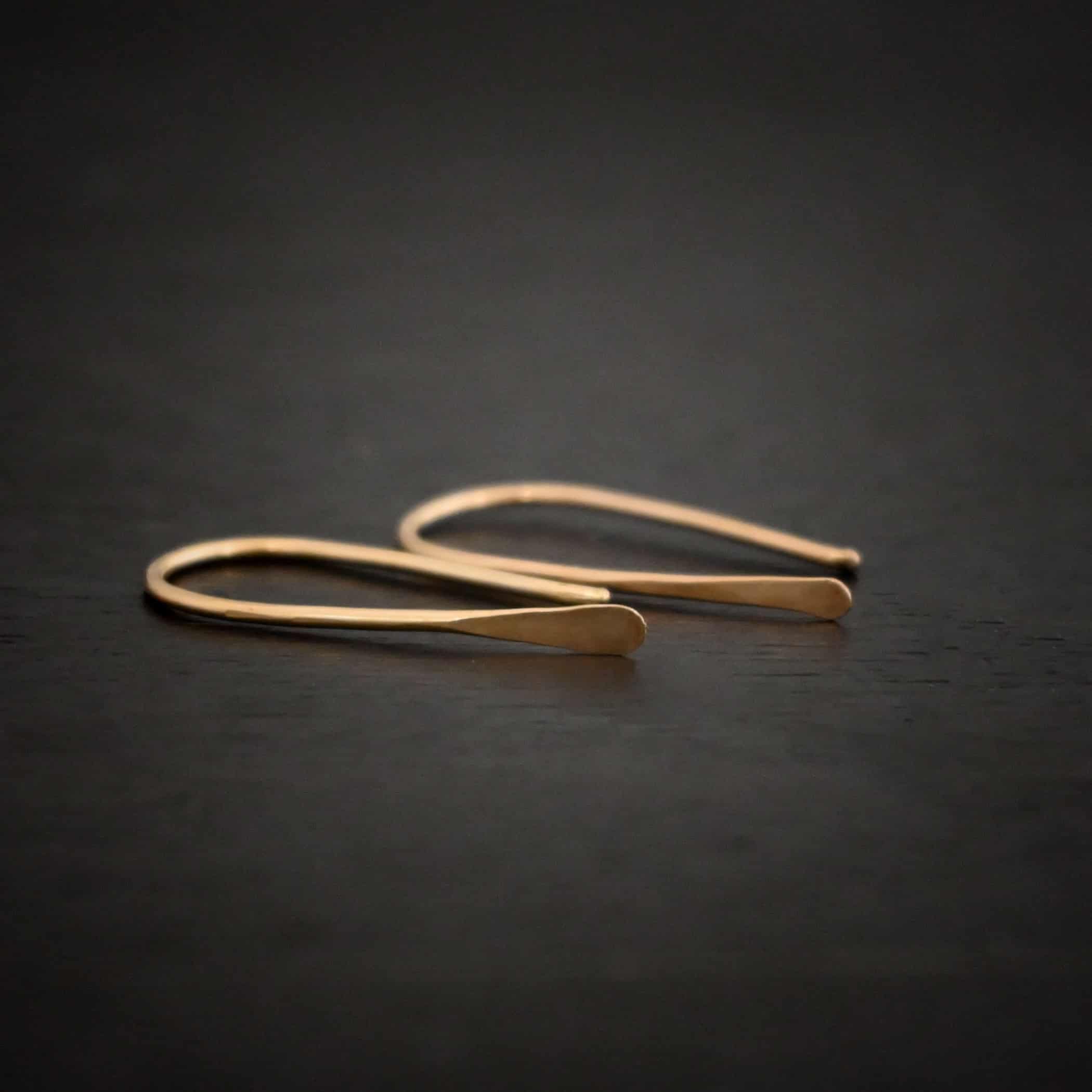 Roadies Photo of the "Roadie" Earrings. Made out of 14k gold fill. Earrings are very simple. They are an ear wire with one end lightly hammered and tipped and the other end of the earring is a hammered paddle. Earrings are lying against a dark background.