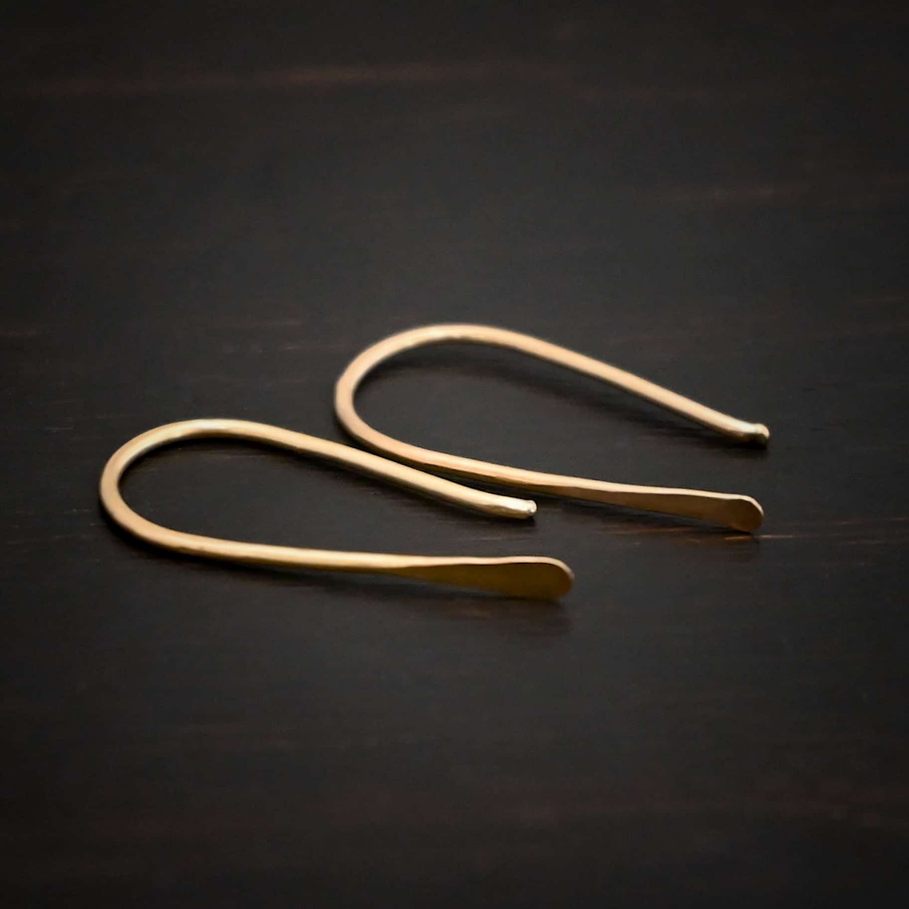 Roadies Photo of the "Roadie" Earrings. Made out of 14k gold. Earrings are very simple. They are an ear wire with one end lightly hammered and tipped and the other end of the earring is a hammered paddle. Earrings are lying against a dark background.