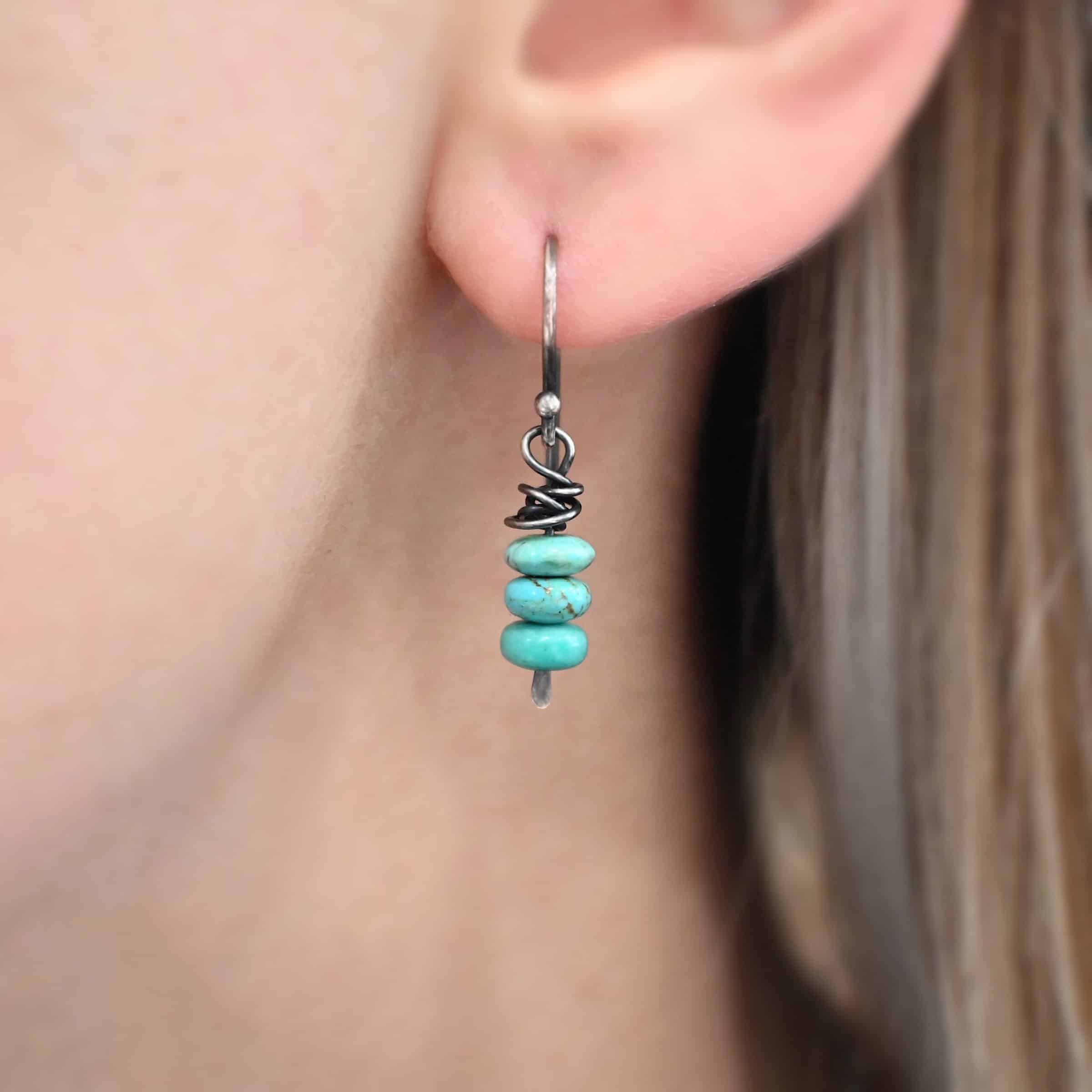 Photo of the "Summer's Breeze" Earring as shown on a model. Made out of sterling silver and turquoise. Earrings have french hook ear wires. Attached to the ear wires on the bottom is a small silver stick with a loop on the top, silver wrapped around the top of the stick after the loop, and a hammered paddle bottom. In between the wrapped silver and paddle bottom are three small pieces of turquoise. Earring is shown up close by a model.