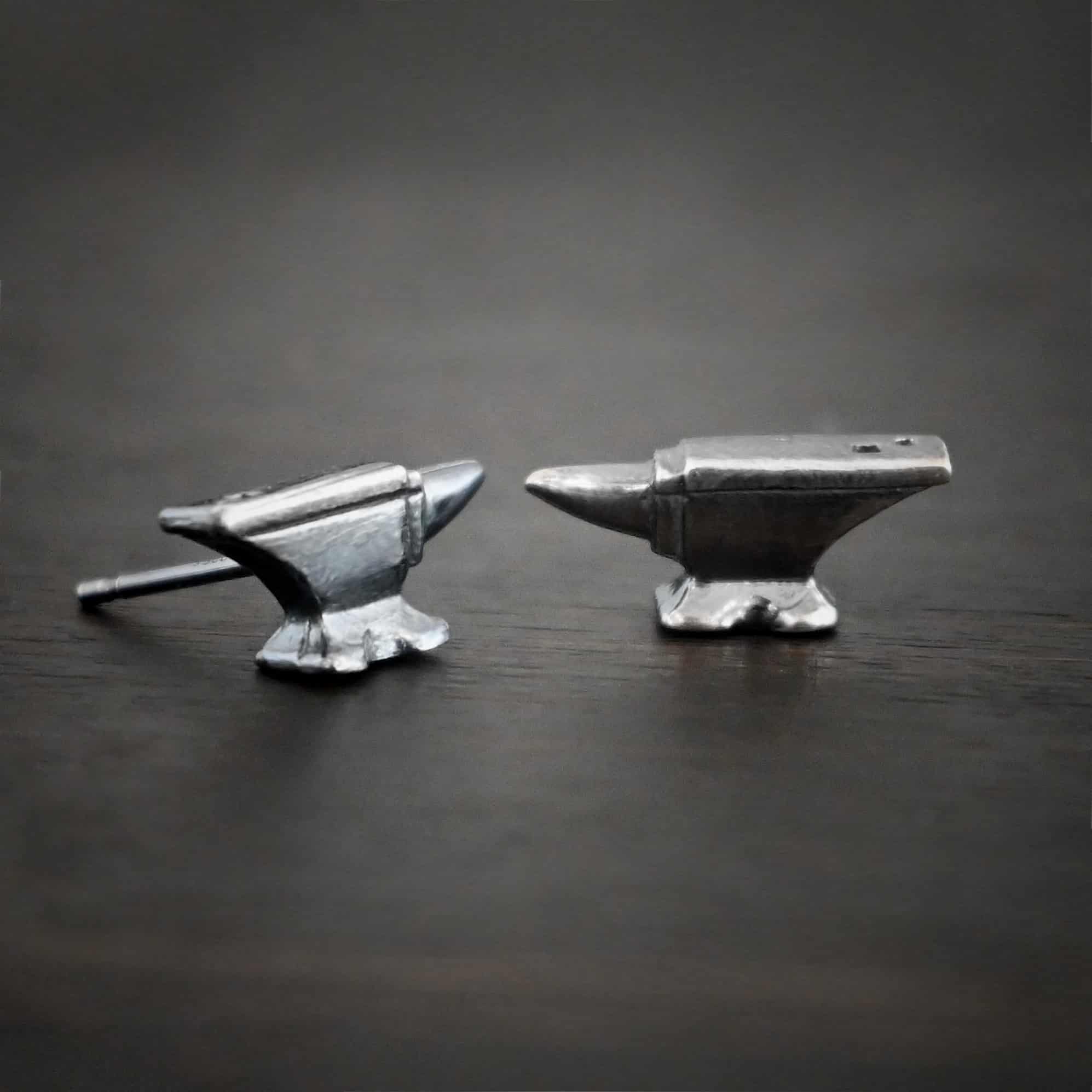 Photo of the "Silversmith" Earrings. Earrings are made out of sterling silver and have post ear wires. Earrings have small anvil charms that the post ear wires are attached to. Earrings are lying against a dark background.