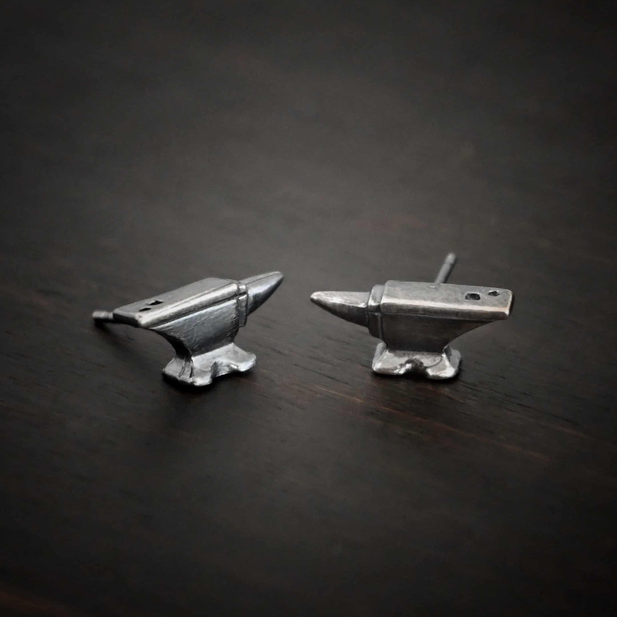 Photo of the "Silversmith" Earrings. Earrings are made out of sterling silver and have post ear wires. Earrings have small anvil charms that the post ear wires are attached to. Earrings are lying against a dark background.
