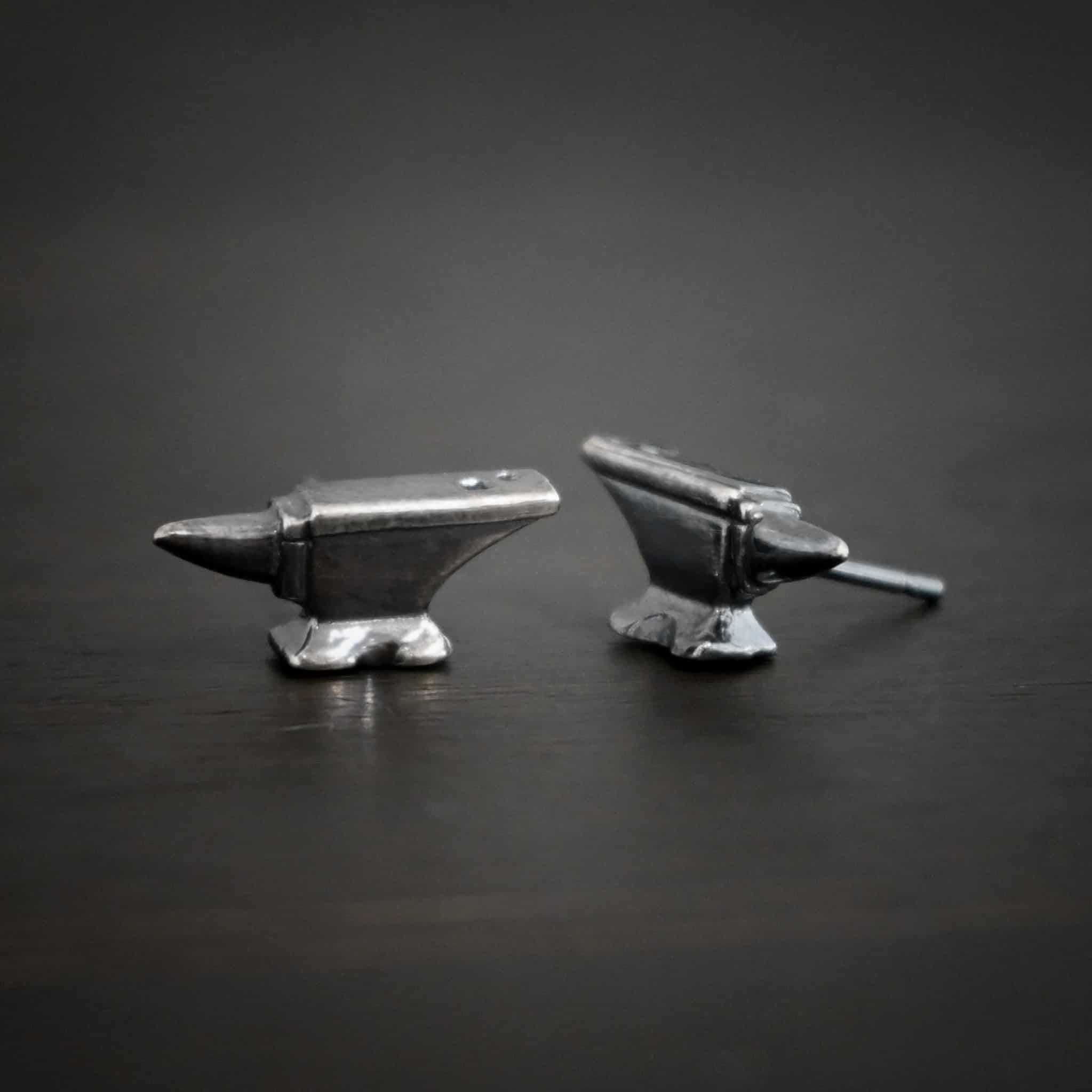Photo of the "Silversmith" Earrings. Earrings are made out of sterling silver and have post ear wires. Earrings have small anvil charms that the post ear wires are attached to. Earrings are lying against a dark background.