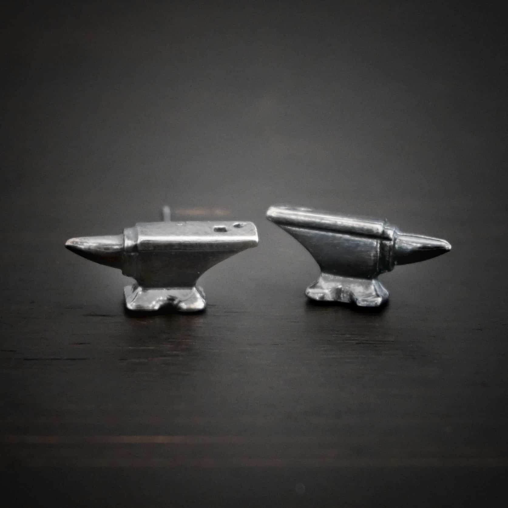 Photo of the "Silversmith" Earrings. Earrings are made out of sterling silver and have post ear wires. Earrings have small anvil charms that the post ear wires are attached to. Earrings are lying against a dark background.