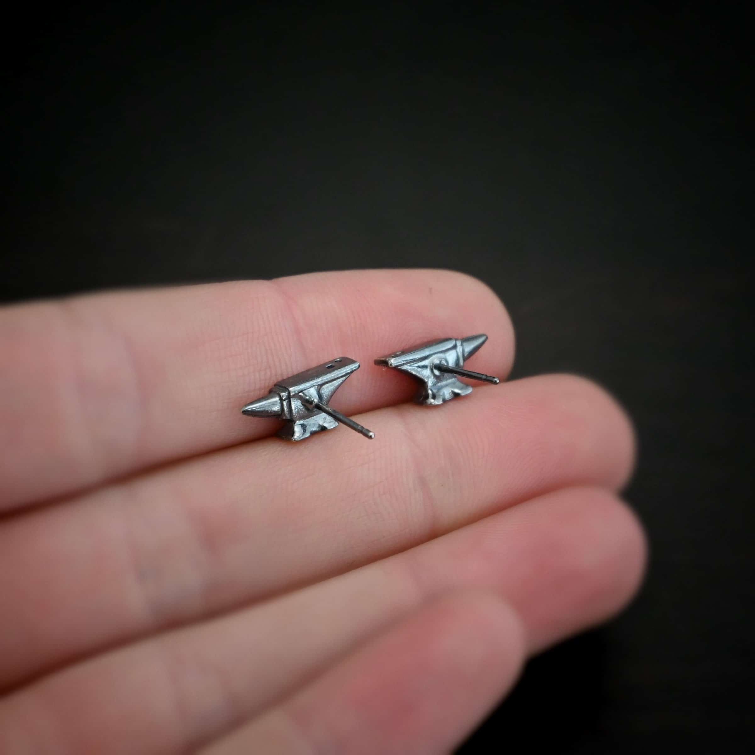 Photo of the "Silversmith" Earrings. Earrings are made out of sterling silver and have post ear wires. Earrings have small anvil charms that the post ear wires are attached to. Earrings are lying on the fingertips of a hand.