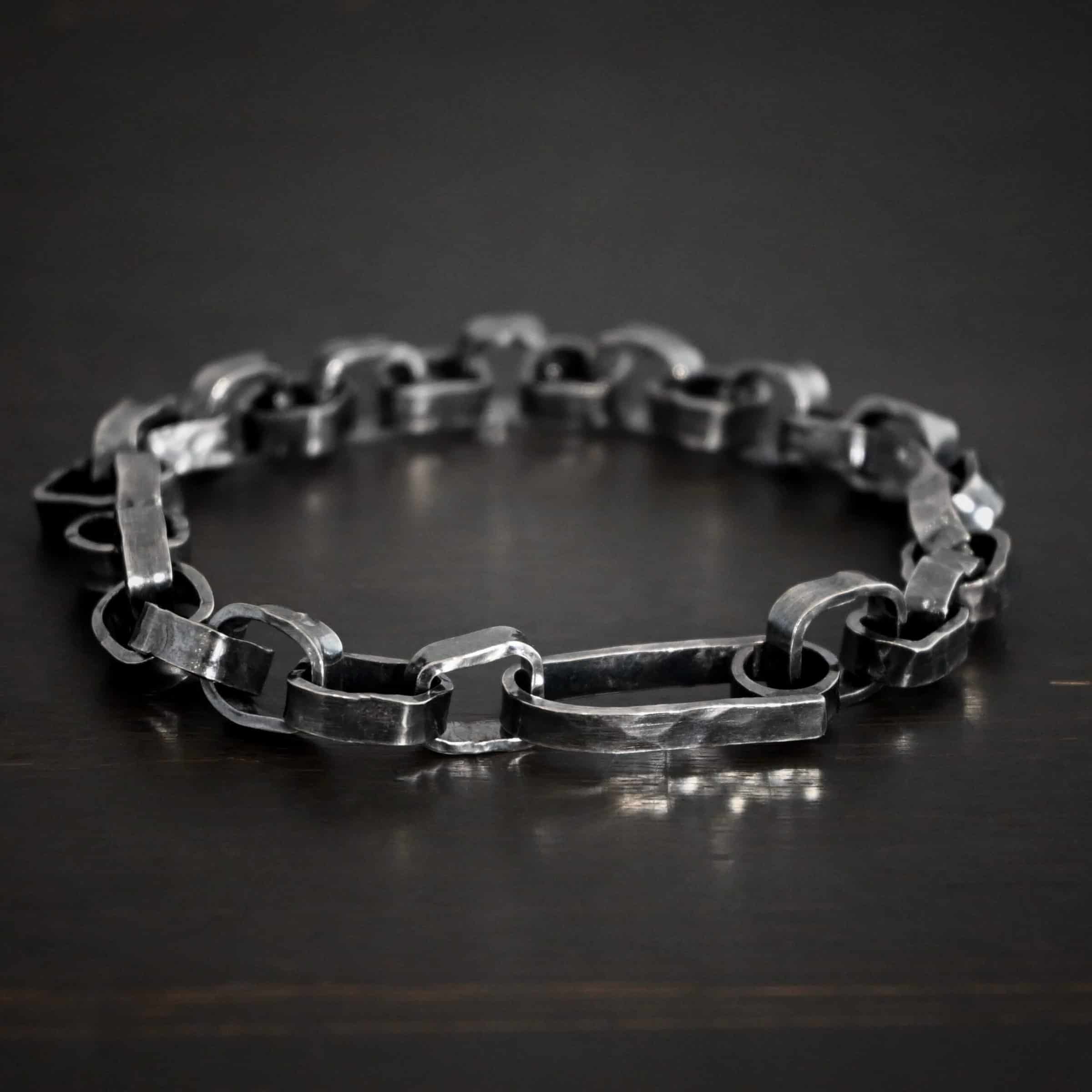 Rustic Bracelet Photo of the "Rustic" Bracelet. Bracelet is made out of sterling silver. Each link is a thick circle link that is hammered and oxidized. The clasp of the bracelet is also thick, hammered, and oxidized, but it is a long oval shape. Bracelet is lying against a dark background.
