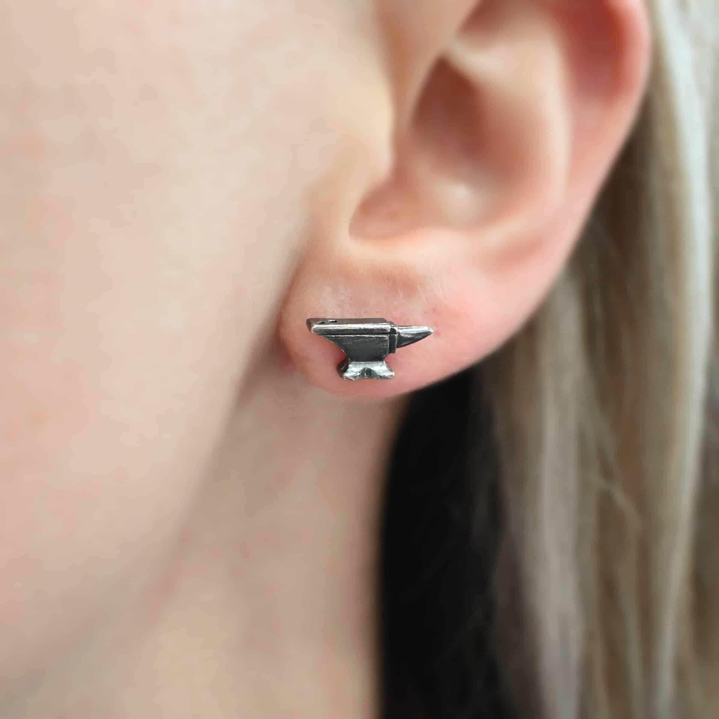 Photo of the "Silversmith" Earring shown on a model. Earring is made out of sterling silver and has a post ear wire. Earring has a small anvil charm that the post ear wire attaches to.