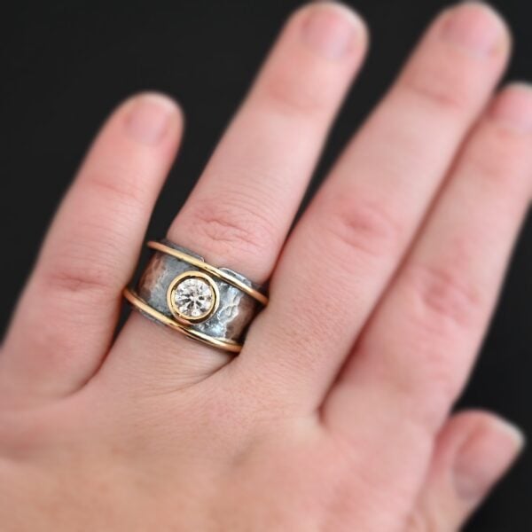 Photo of the "Banded Together" Ring. Made out of sterling silver, gold, and moissanite. The ring band does not fully connect. It is thick and hammered. There is a moissanite in the center that connects both sides. It is set in gold. There is a thin band of gold on top and on the bottom of the ring. They both go fully around and connect both sides of the ring as well. Ring is worn by a model.