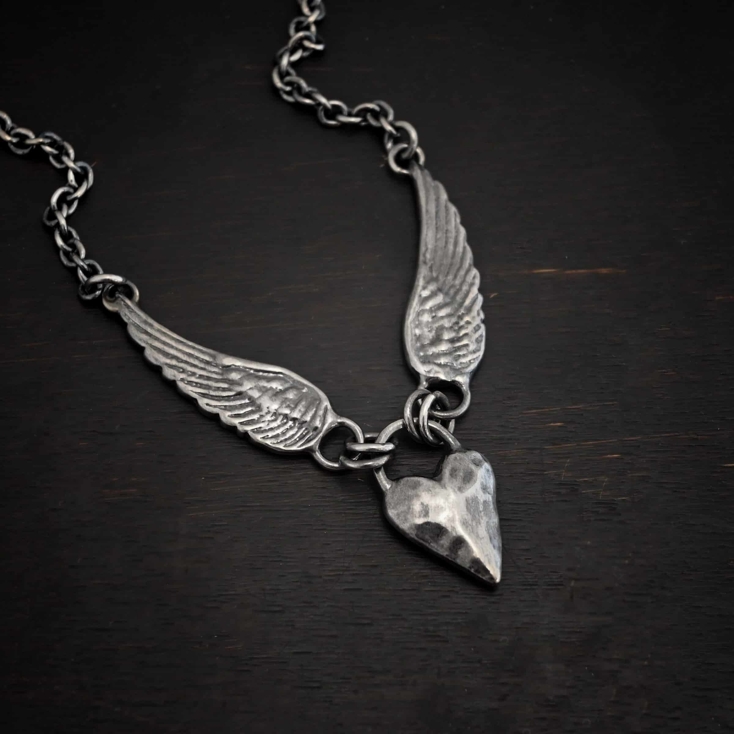 Courage Necklace Photo of the "Courage" Necklace. Made out of sterling silver. There is a thick, solid sterling silver heart that is a medium size for the pendant. It is hammered and oxidized. There is a thick semicircle that is soldered on both sides of the heart on the top. There are four jump rings attached to the semicircle, two on each side. Attached to the jump rings on each side are two angel wings that face opposite directions. The tip of each wing has a loop soldered to the back of them. Attached to the loop on the back of each wing is the chain of the necklace. Necklace is lying against a dark background.