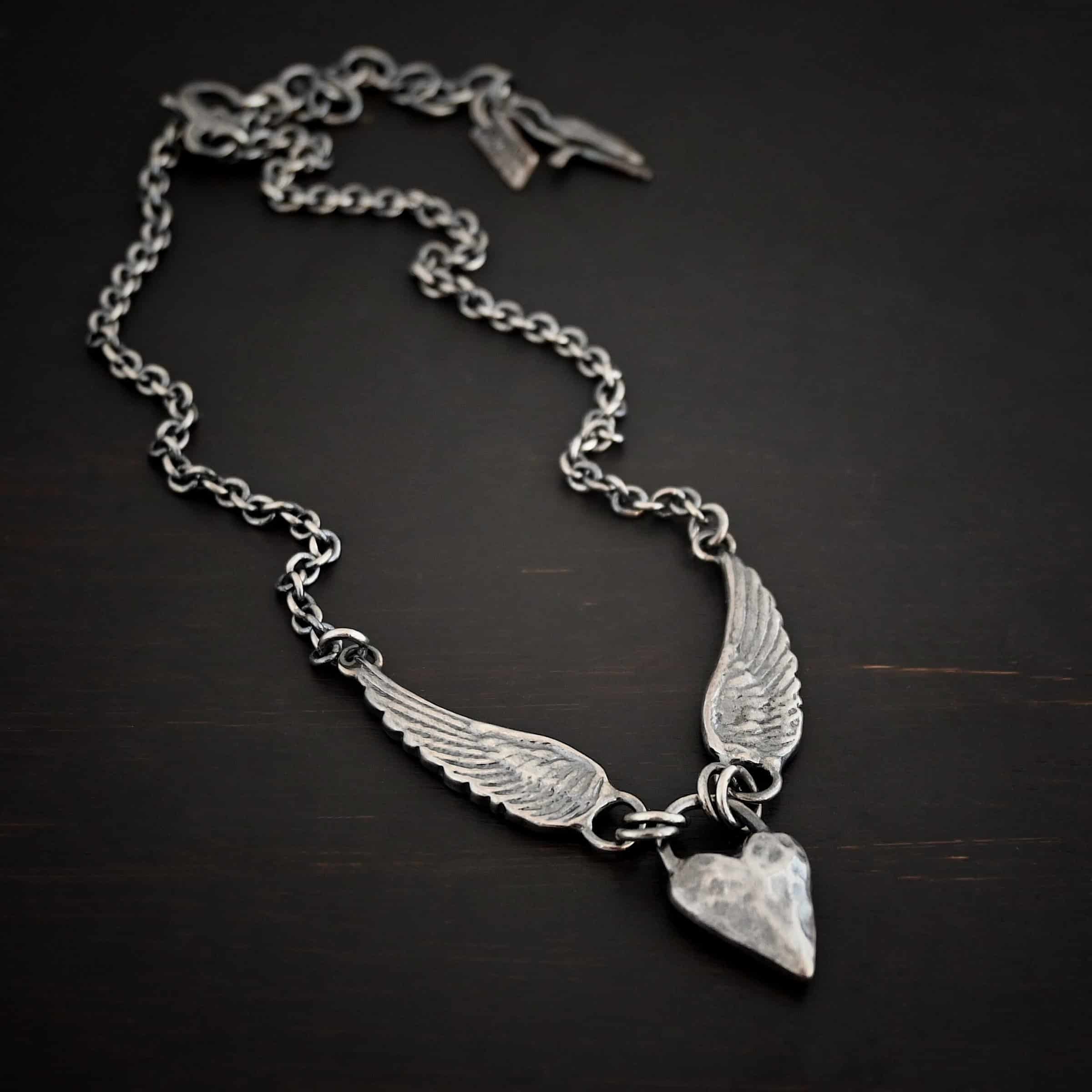Courage Necklace Photo of the "Courage" Necklace. Made out of sterling silver. There is a thick, solid sterling silver heart that is a medium size for the pendant. It is hammered and oxidized. There is a thick semicircle that is soldered on both sides of the heart on the top. There are four jump rings attached to the semicircle, two on each side. Attached to the jump rings on each side are two angel wings that face opposite directions. The tip of each wing has a loop soldered to the back of them. Attached to the loop on the back of each wing is the chain of the necklace. Necklace is lying against a dark background.