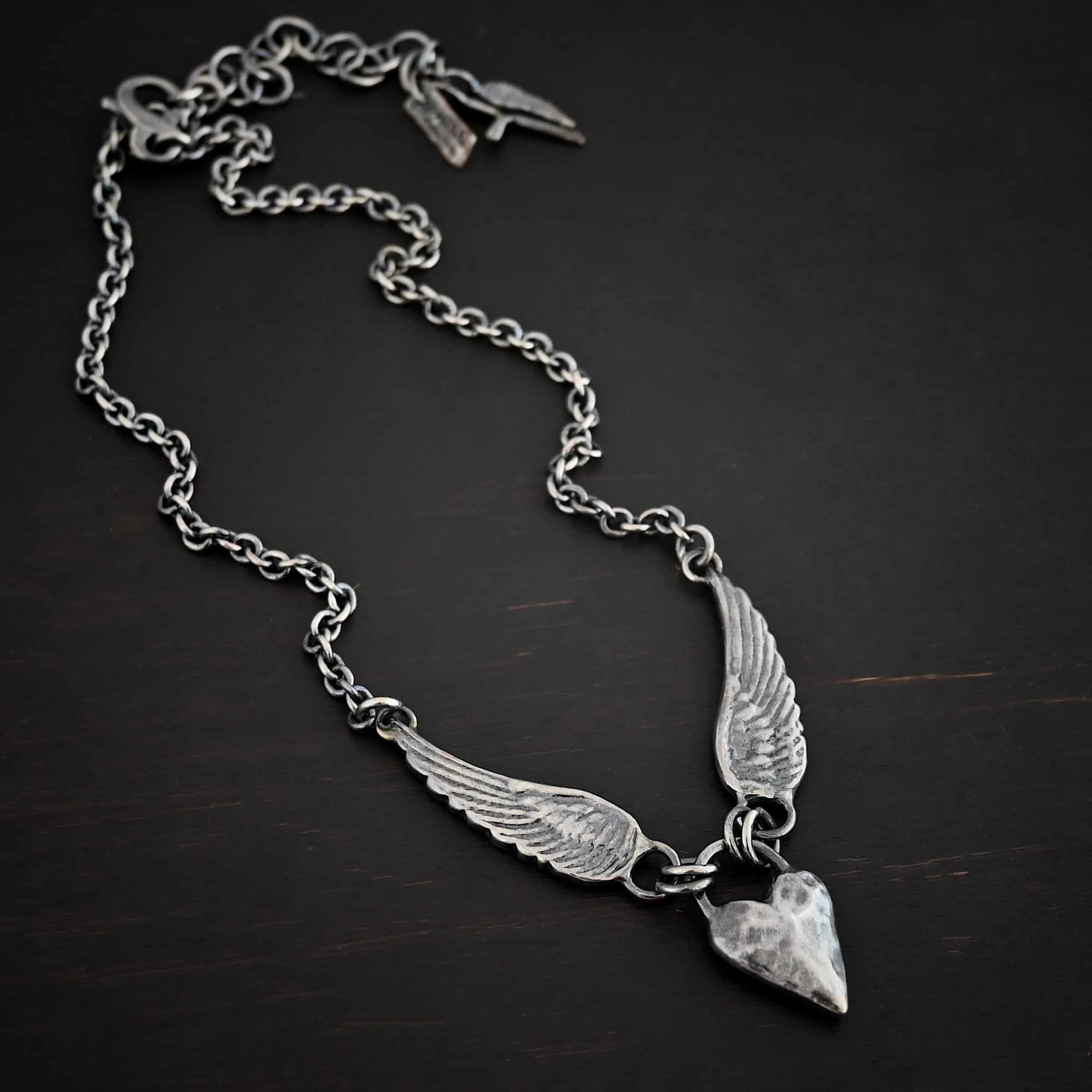 Courage Necklace Photo of the "Courage" Necklace. Made out of sterling silver. There is a thick, solid sterling silver heart that is a medium size for the pendant. It is hammered and oxidized. There is a thick semicircle that is soldered on both sides of the heart on the top. There are four jump rings attached to the semicircle, two on each side. Attached to the jump rings on each side are two angel wings that face opposite directions. The tip of each wing has a loop soldered to the back of them. Attached to the loop on the back of each wing is the chain of the necklace. Necklace is lying against a dark background.