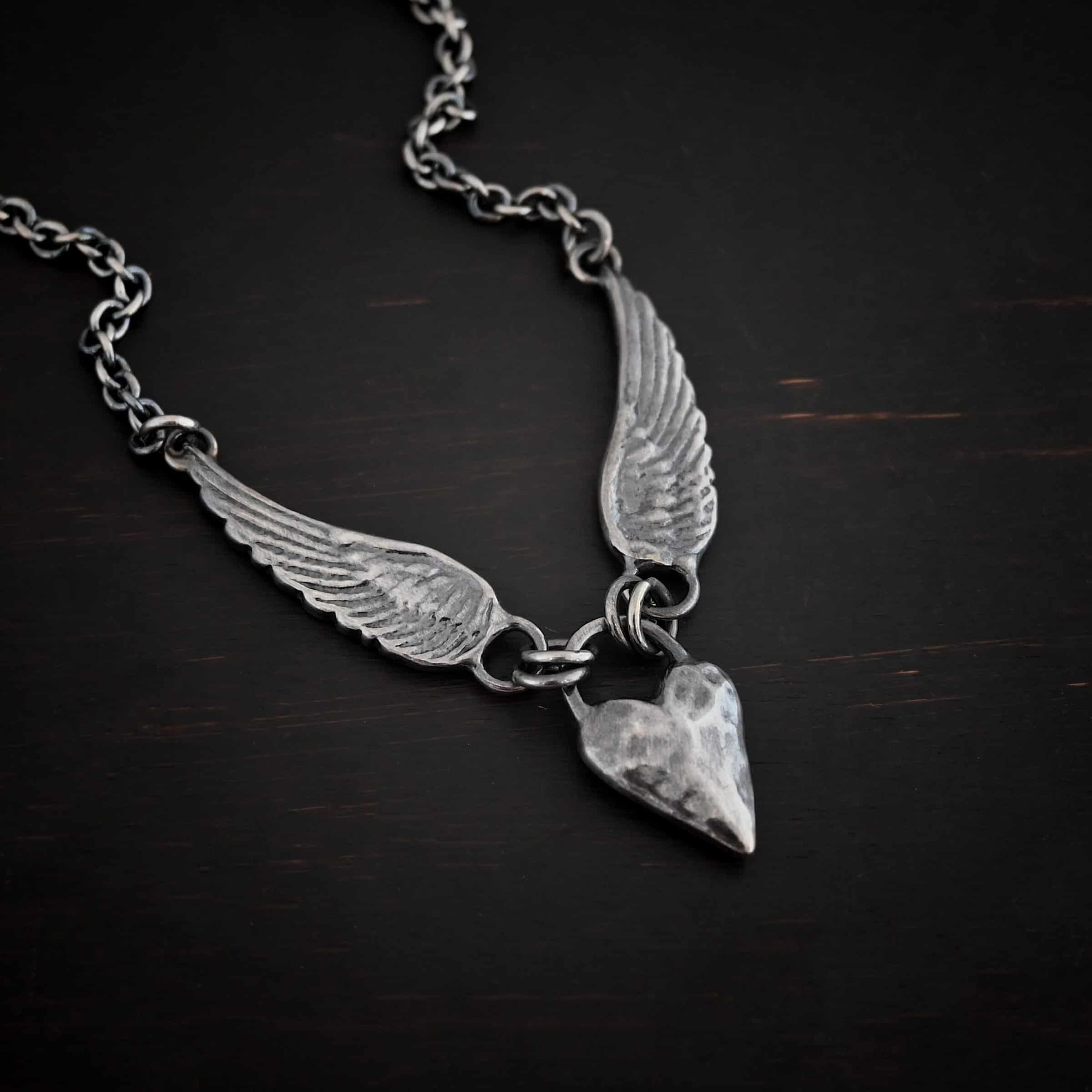 Courage Necklace Photo of the "Courage" Necklace. Made out of sterling silver. There is a thick, solid sterling silver heart that is a medium size for the pendant. It is hammered and oxidized. There is a thick semicircle that is soldered on both sides of the heart on the top. There are four jump rings attached to the semicircle, two on each side. Attached to the jump rings on each side are two angel wings that face opposite directions. The tip of each wing has a loop soldered to the back of them. Attached to the loop on the back of each wing is the chain of the necklace. Necklace is lying against a dark background.