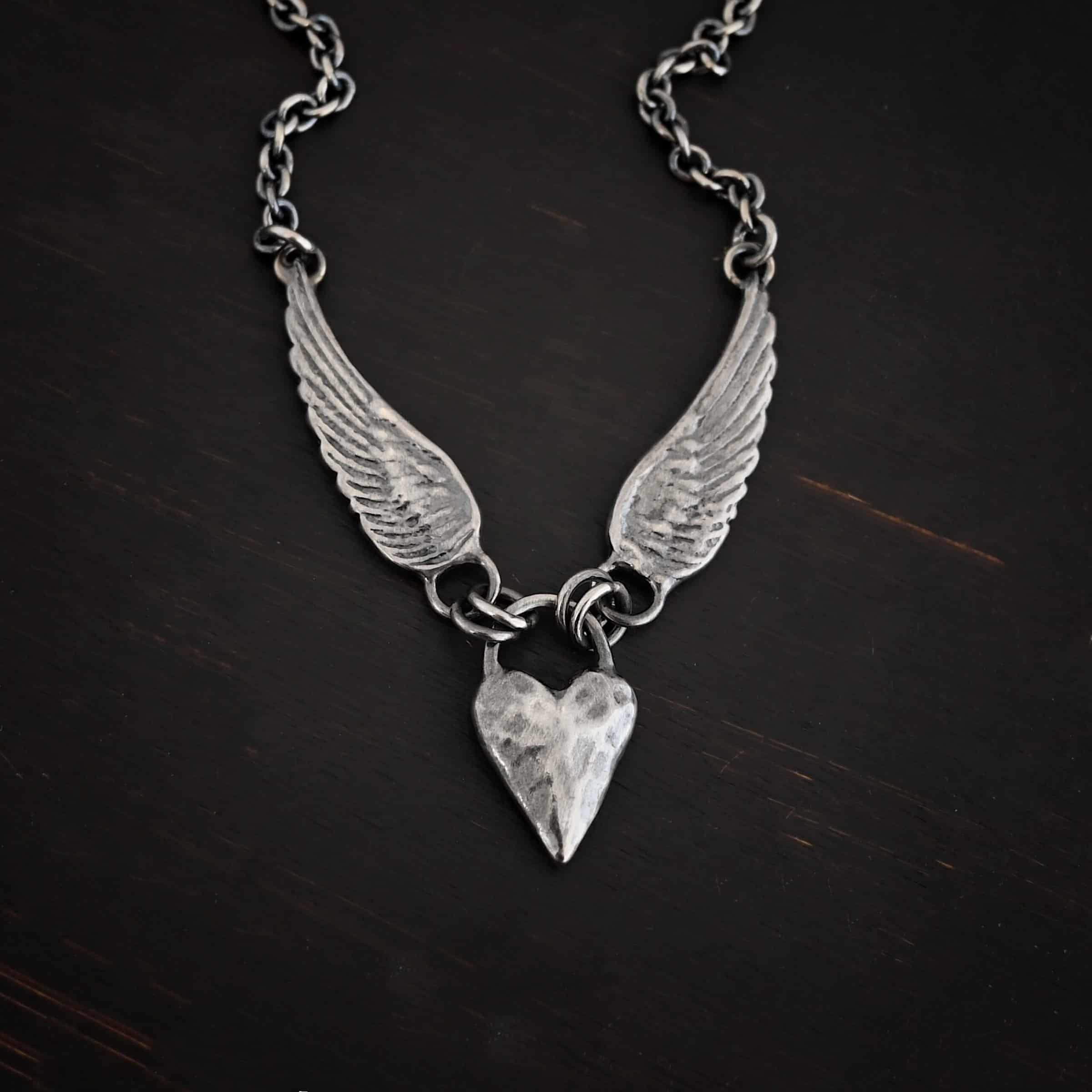 Courage Necklace Photo of the "Courage" Necklace. Made out of sterling silver. There is a thick, solid sterling silver heart that is a medium size for the pendant. It is hammered and oxidized. There is a thick semicircle that is soldered on both sides of the heart on the top. There are four jump rings attached to the semicircle, two on each side. Attached to the jump rings on each side are two angel wings that face opposite directions. The tip of each wing has a loop soldered to the back of them. Attached to the loop on the back of each wing is the chain of the necklace. Necklace is lying against a dark background.